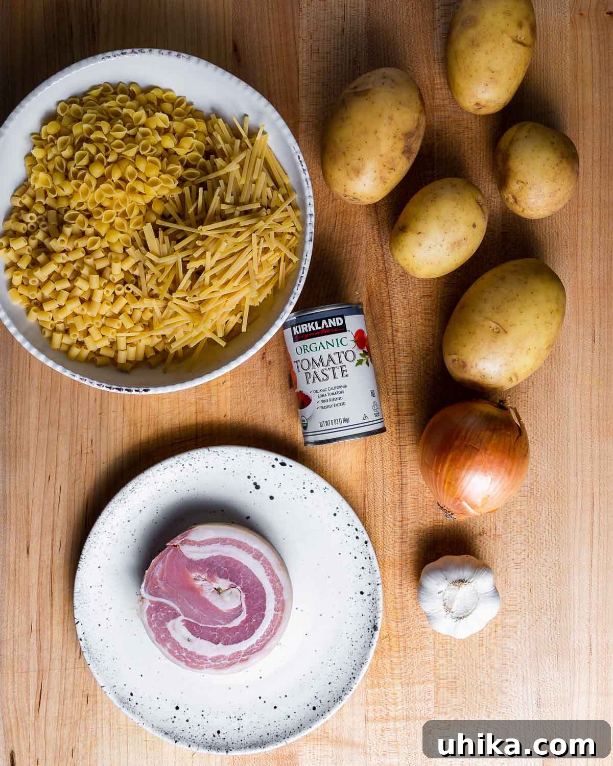 Pasta with Potatoes 4 A flat-lay photograph showcasing the fresh, wholesome ingredients required for Pasta e Patate, including a bowl of assorted small pasta, whole and diced Yukon Gold potatoes, rich tomato paste, savory pancetta, a fragrant onion, and cloves of garlic.