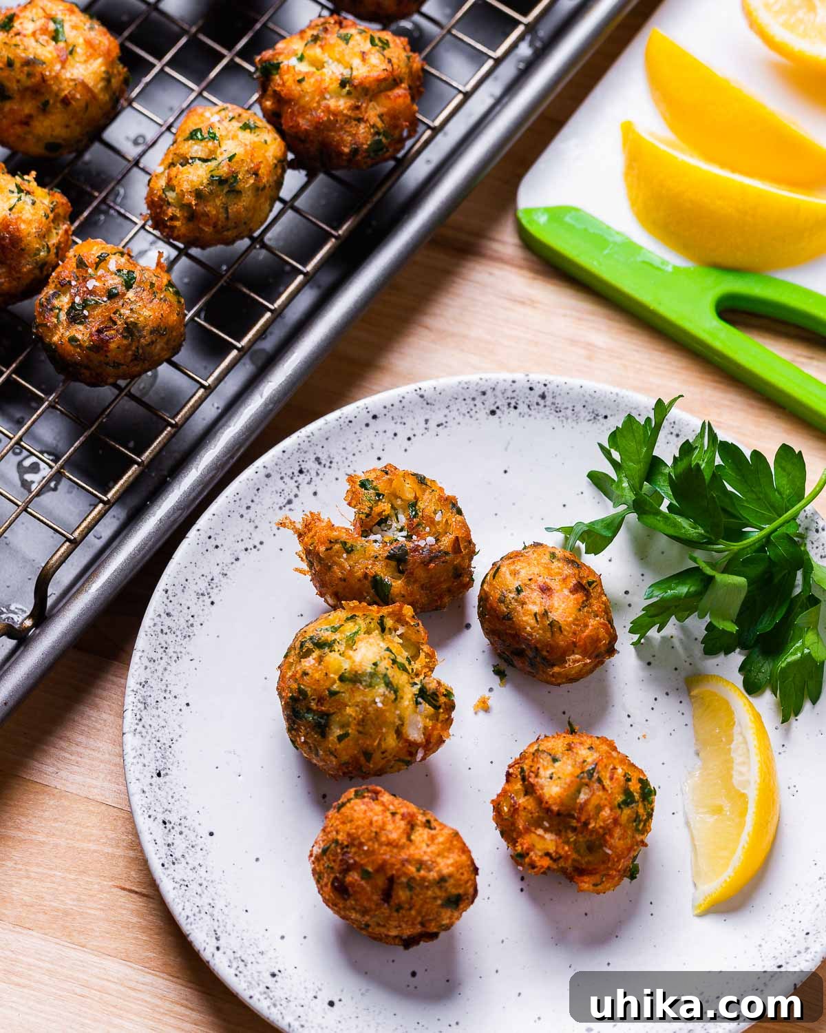 Mediterranean Salt Cod Fritters 10 Baccala fritters arranged beautifully on a white plate and some draining on a wire rack, with fresh lemon wedges on a wooden cutting board.