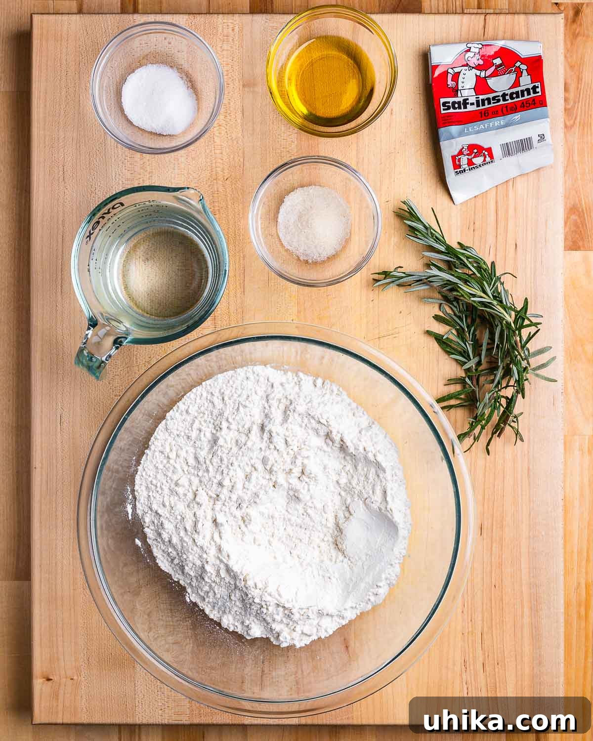 Effortless No-Knead Focaccia 3 Neatly arranged ingredients for no-knead focaccia: SAF instant yeast, olive oil, salt, sugar, water, fresh rosemary, and bread flour.