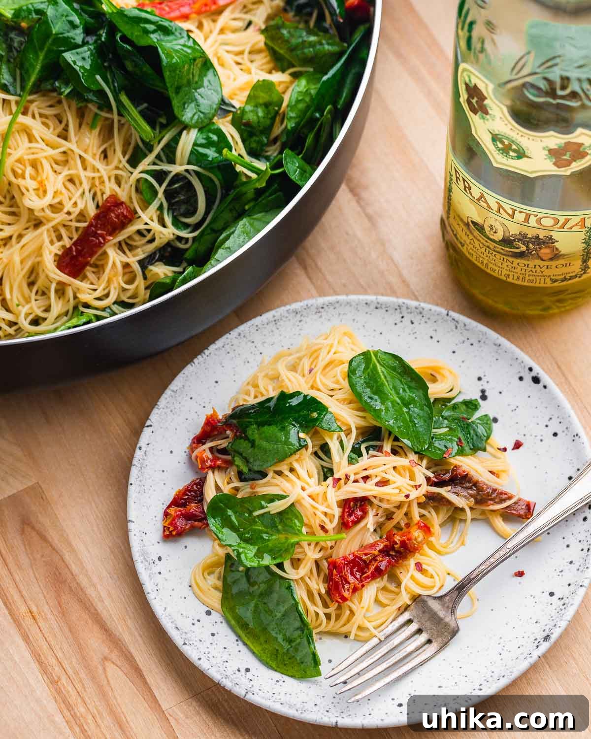 Sun-Drenched Spinach and Tomato Pasta 7 White plate with spinach sun dried tomato pasta along with olive oil and large pan in background, showcasing a finished dish.