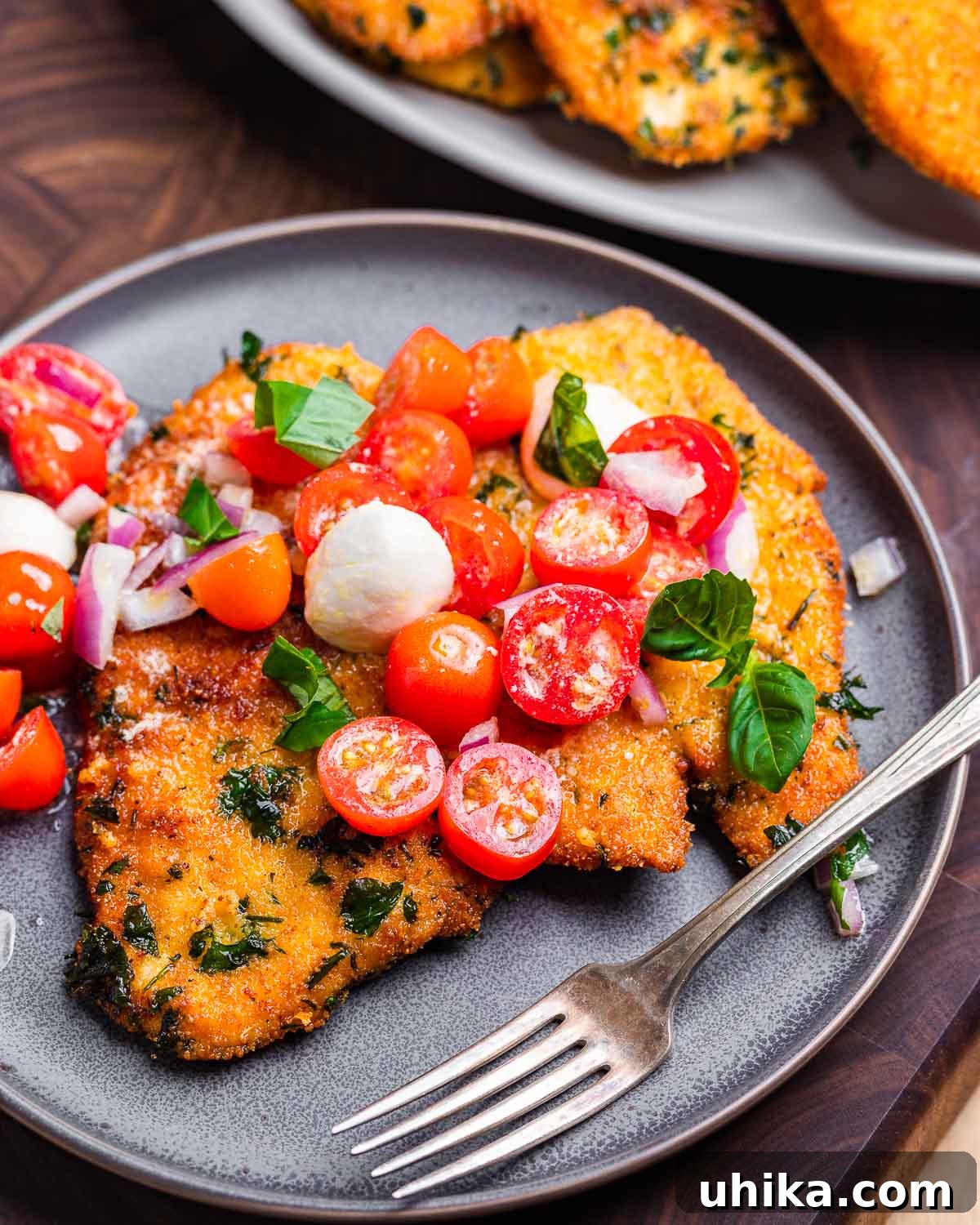 Caprese Chicken Milanese 2 Golden-brown chicken Milanese cutlet on a grey plate, generously topped with a vibrant salad of halved cherry tomatoes, fresh mozzarella balls, and green basil leaves, lightly dressed with olive oil.