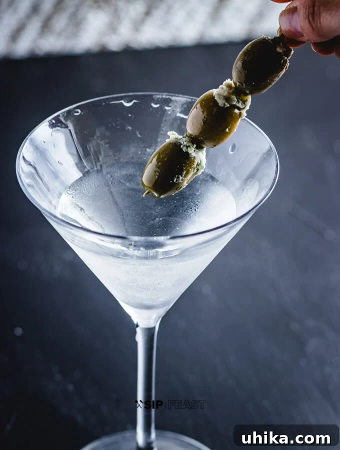 Blue Cheese Olive Martini - sipandfeast.com #martini #cocktails #bluecheese #olives Three plump blue cheese stuffed olives being carefully placed into a freshly poured gin martini with a cocktail pick, adding a gourmet touch.