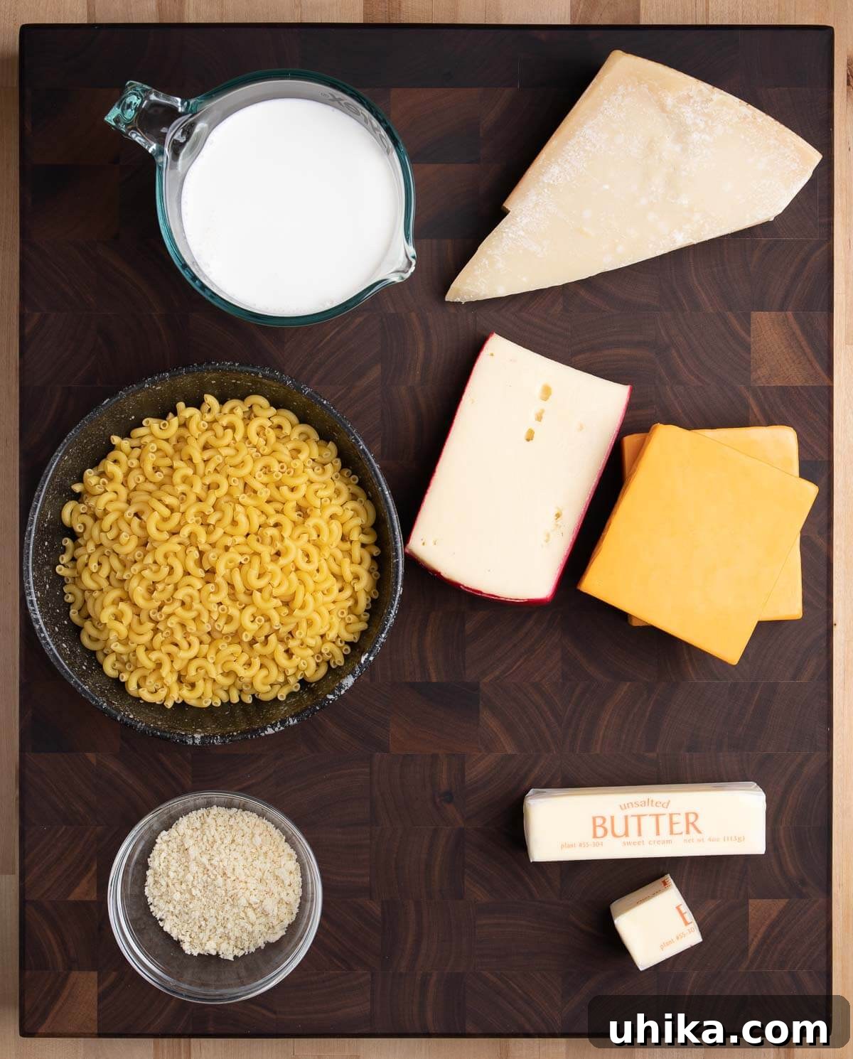 Golden Crusted Creamy Macaroni Bake 3 Ingredients for baked macaroni and cheese displayed on a table: milk, Parmigiano Reggiano, elbow macaroni, Fontina, cheddar, panko breadcrumbs, and butter.