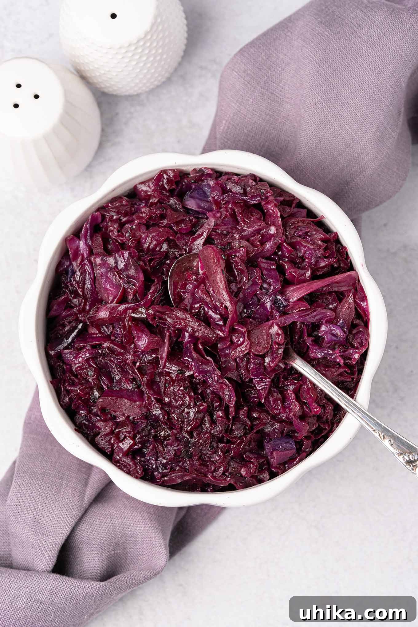 Sweet and Tangy Braised Red Cabbage 2 An overhead view of a serving bowl of braised red cabbage with a spoon in it, on top of a linen napkin with white ceramic salt and pepper shakers behind it and to the left.