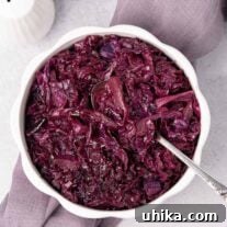 An overhead view of a serving bowl of braised red cabbage with a spoon in it, on top of a linen napkin with white ceramic salt and pepper shakers behind it and to the left.