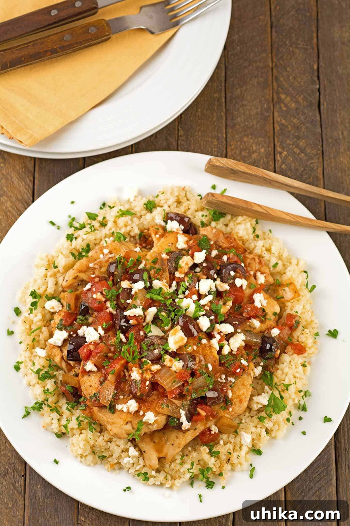 Wholesome Mediterranean Chicken and Cauliflower Rice 2 A dinner plate with boneless chicken breasts, topped with a light sauce of tomatoes, Kalamata olives and feta cheese served over a bed of riced cauliflower, extra plates and napkins in the background.