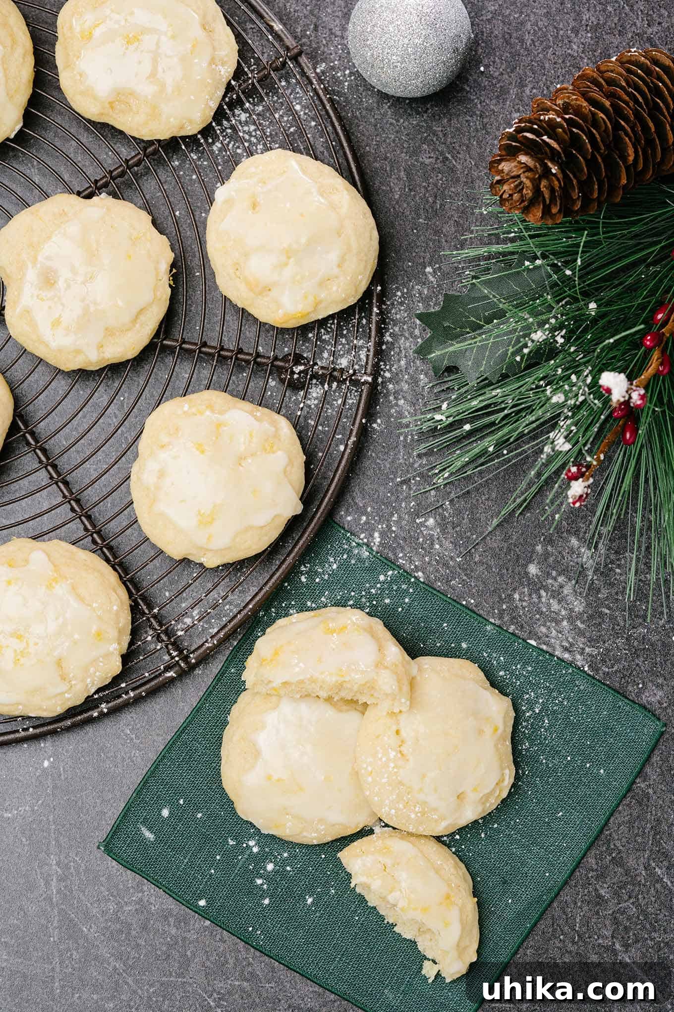 Pillowy Lemon Ricotta Delights 2 An overhead view of three lemon ricotta cookies glazed and sprinkled with powdered sugar on a linen napkin, a cooling rack with more cookies to the left.