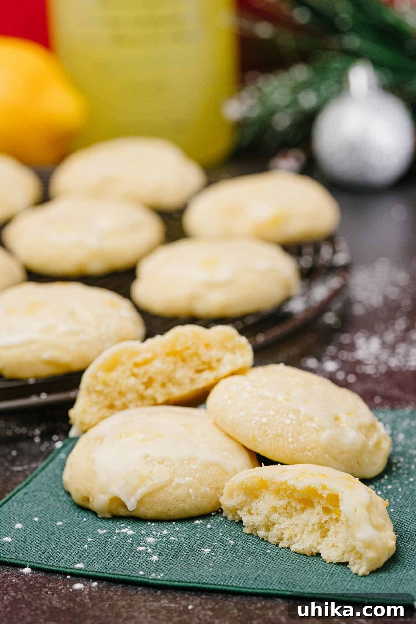 Pillowy Lemon Ricotta Delights 3 A serving of lemon ricotta cookies on a green napkin with more cookies, a bottle of limoncello, and Christmas table decor in the background.