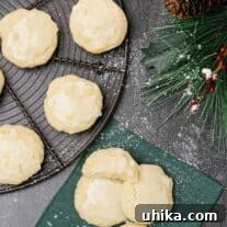 Pillowy Lemon Ricotta Delights 4 An overhead view of three lemon ricotta cookies glazed and sprinkled with powdered sugar on a linen napkin, a cooling rack with more cookies to the left.