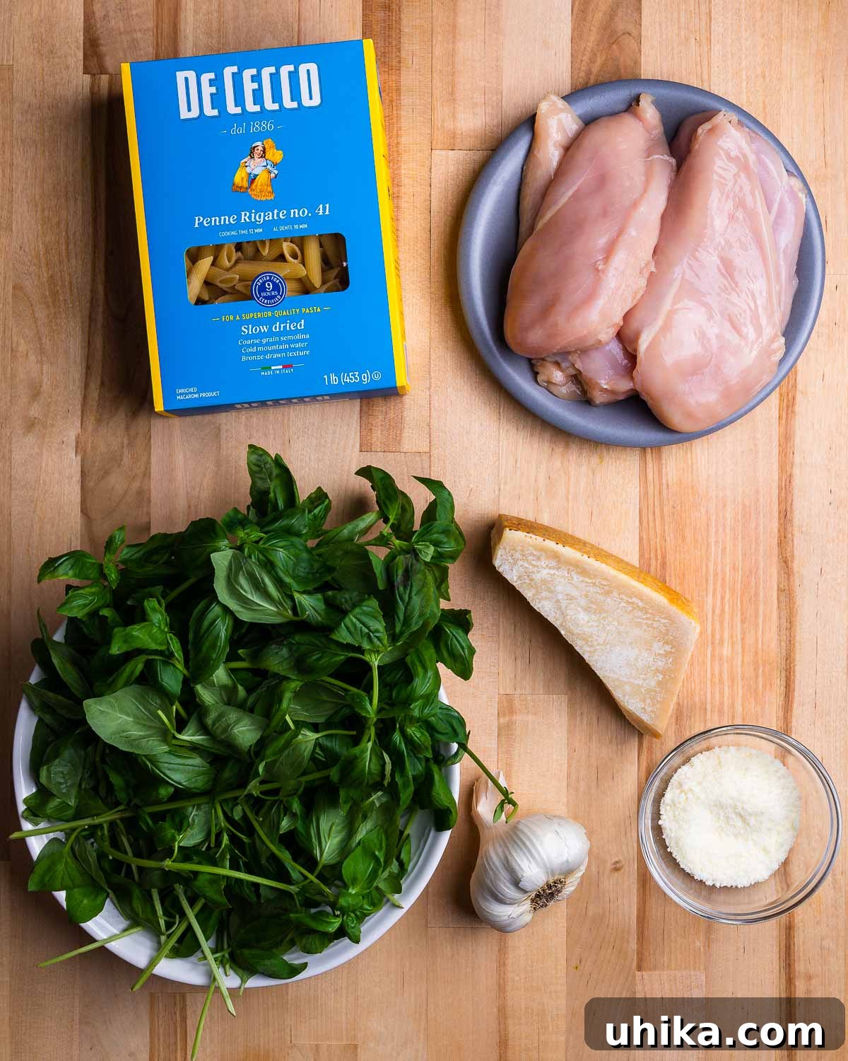 Basil Pesto Chicken Pasta 3 Ingredients for chicken pesto pasta salad shown: penne pasta, chicken breasts, fresh basil, garlic cloves, parmesan cheese, and pecorino cheese laid out on a wooden board.