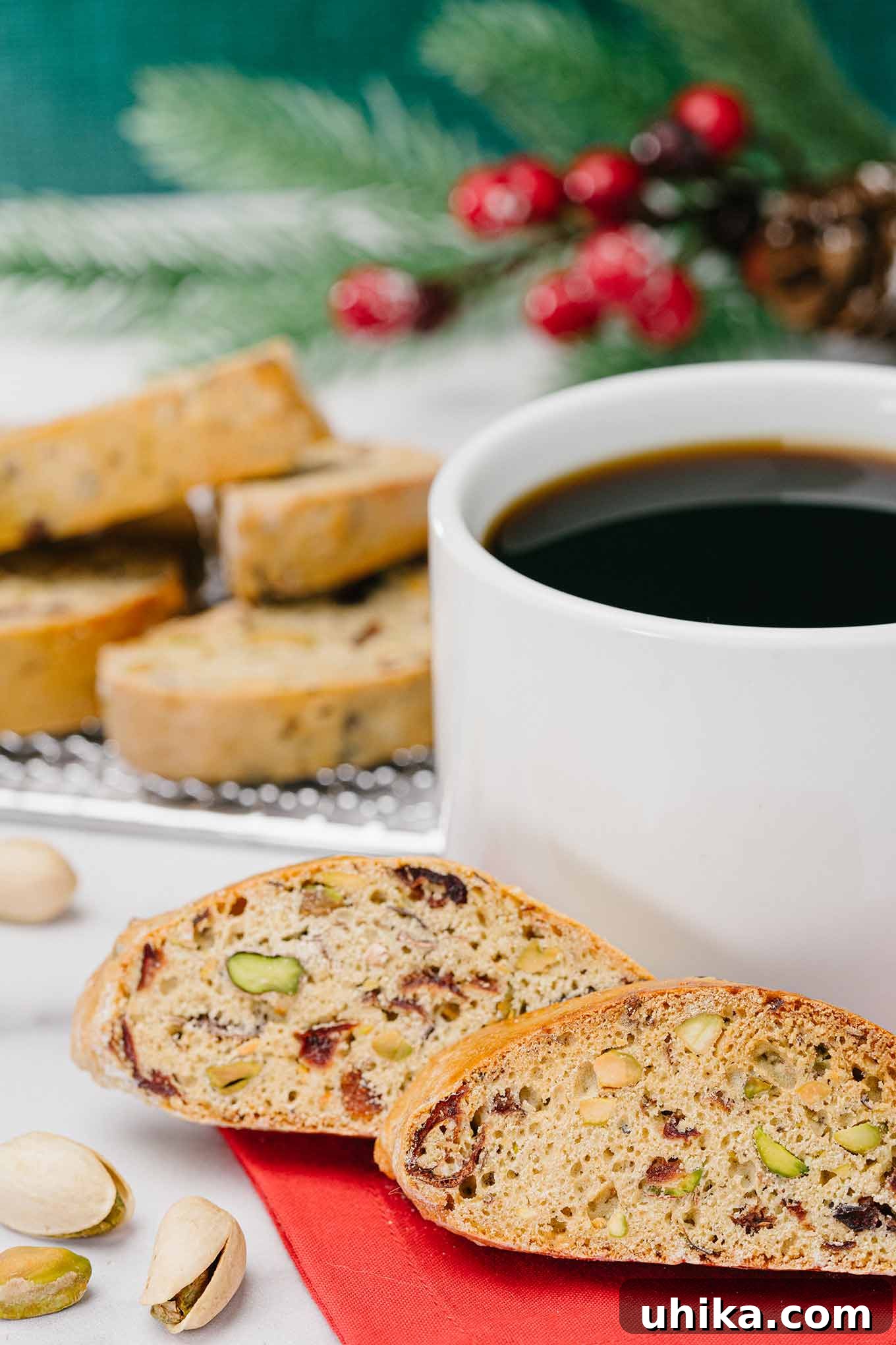 Jeweled Cranberry Pistachio Biscotti 2 Two cranberry pistachio biscotti on a red napkin next to a cup of black coffee with more biscotti in the background.