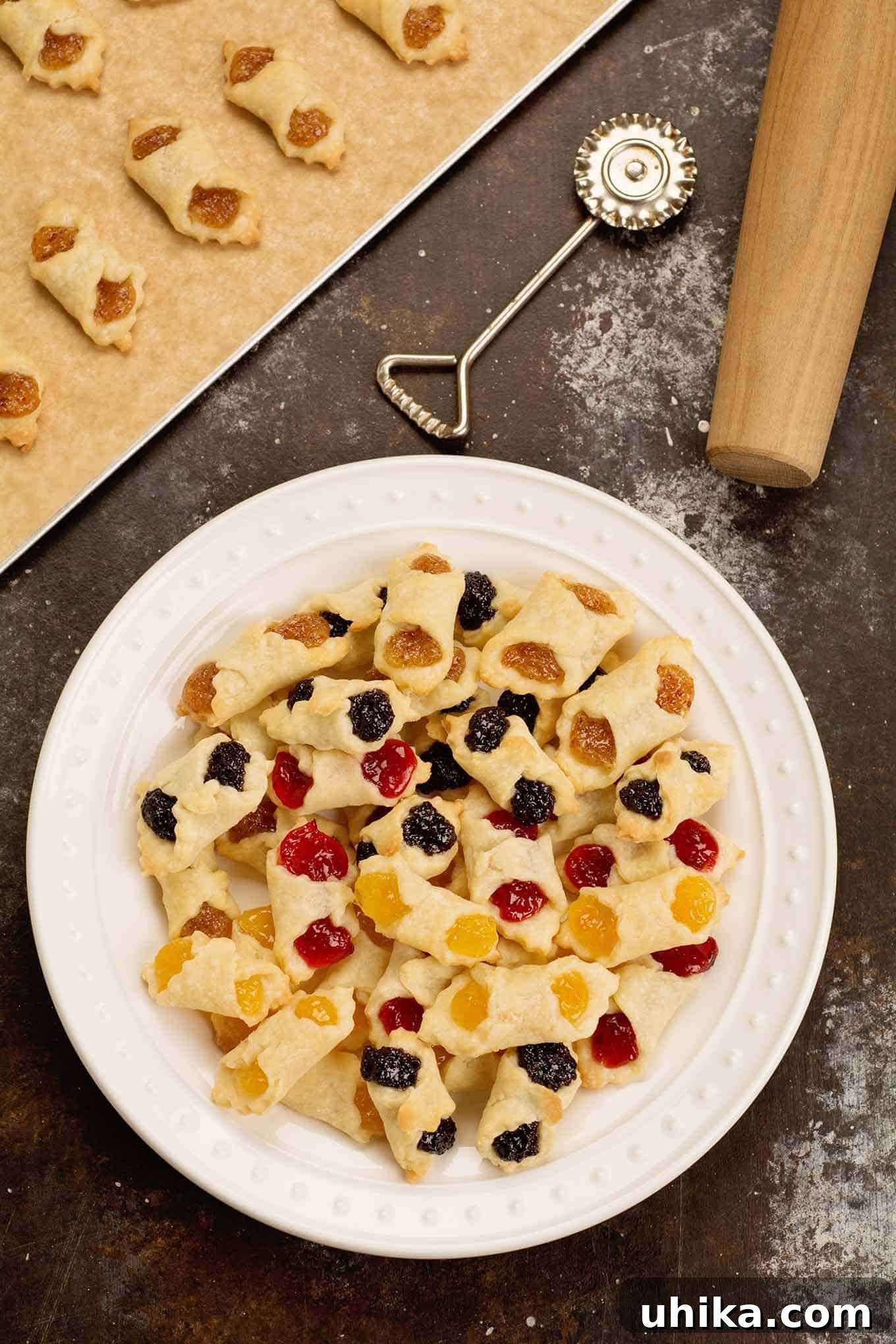 Generations of Hungarian Kiffles 2 A white plate of assorted flavors of Hungarian kiffles with a cookie sheet, pastry wheel, and rolling pin in the background.