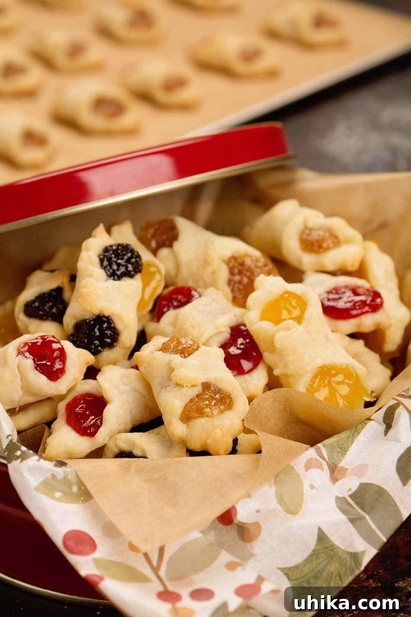 Generations of Hungarian Kiffles 4 A red Christmas gift tin filled with apricot, almond, poppy seed, and raspberry Hungarian kiffles, a baking sheet with more freshly baked kiffles in the background.