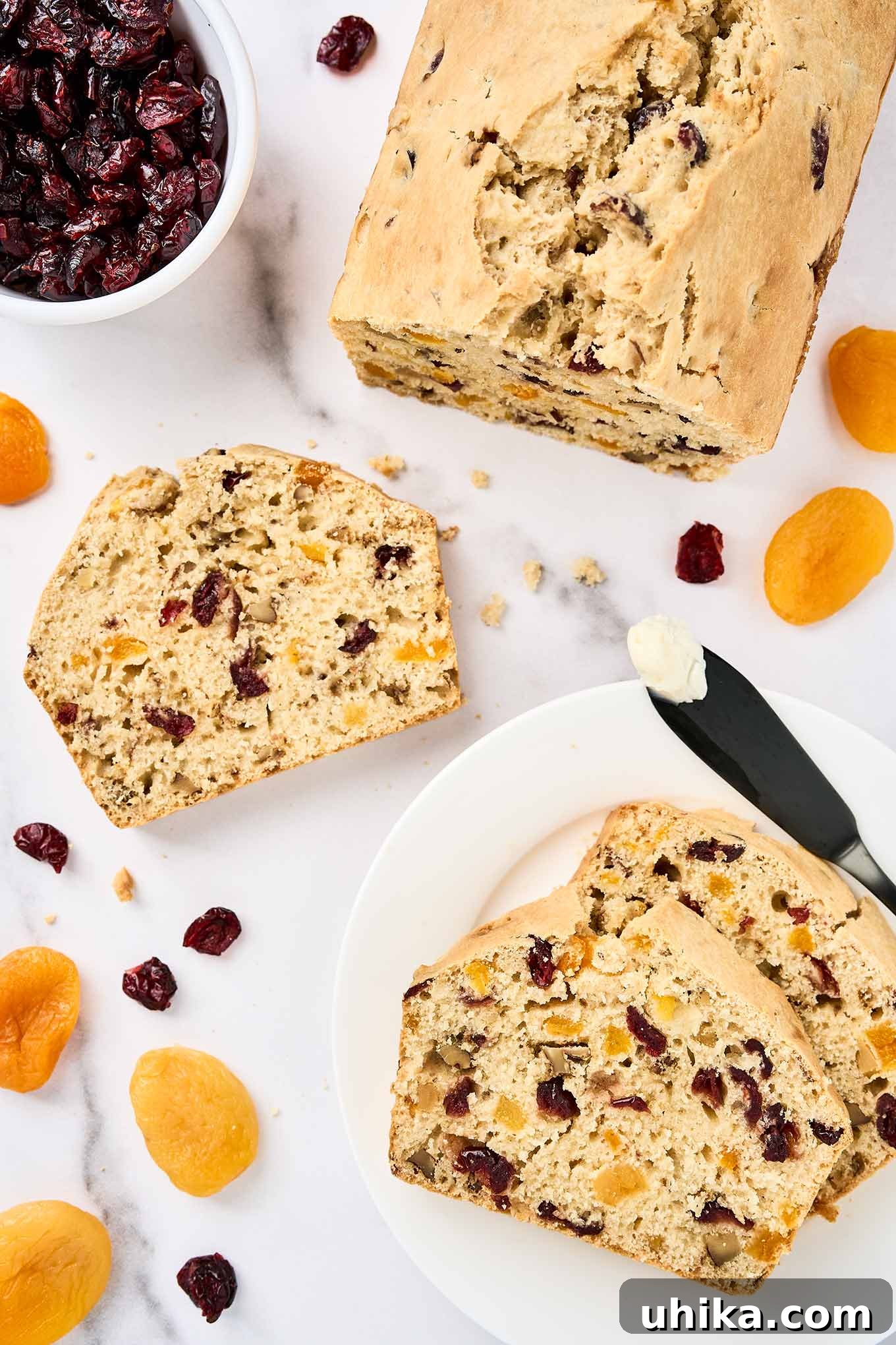 Sun-Kissed Apricot Cranberry Loaf 2 Sliced apricot bread with cranberries and walnuts on a white marble surface, surrounded by dried apricots, cranberries, and a buttered knife.