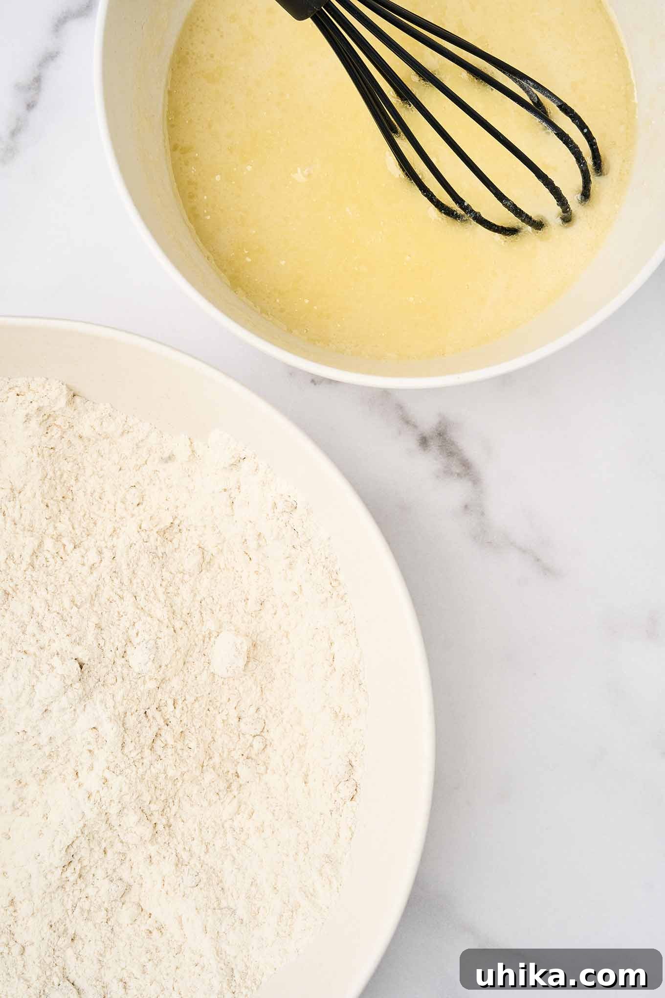 Sun-Kissed Apricot Cranberry Loaf 4 Two mixing bowls side by side: one with flour and dry ingredients, the other with whisked wet ingredients and a black whisk, set on a marble surface.