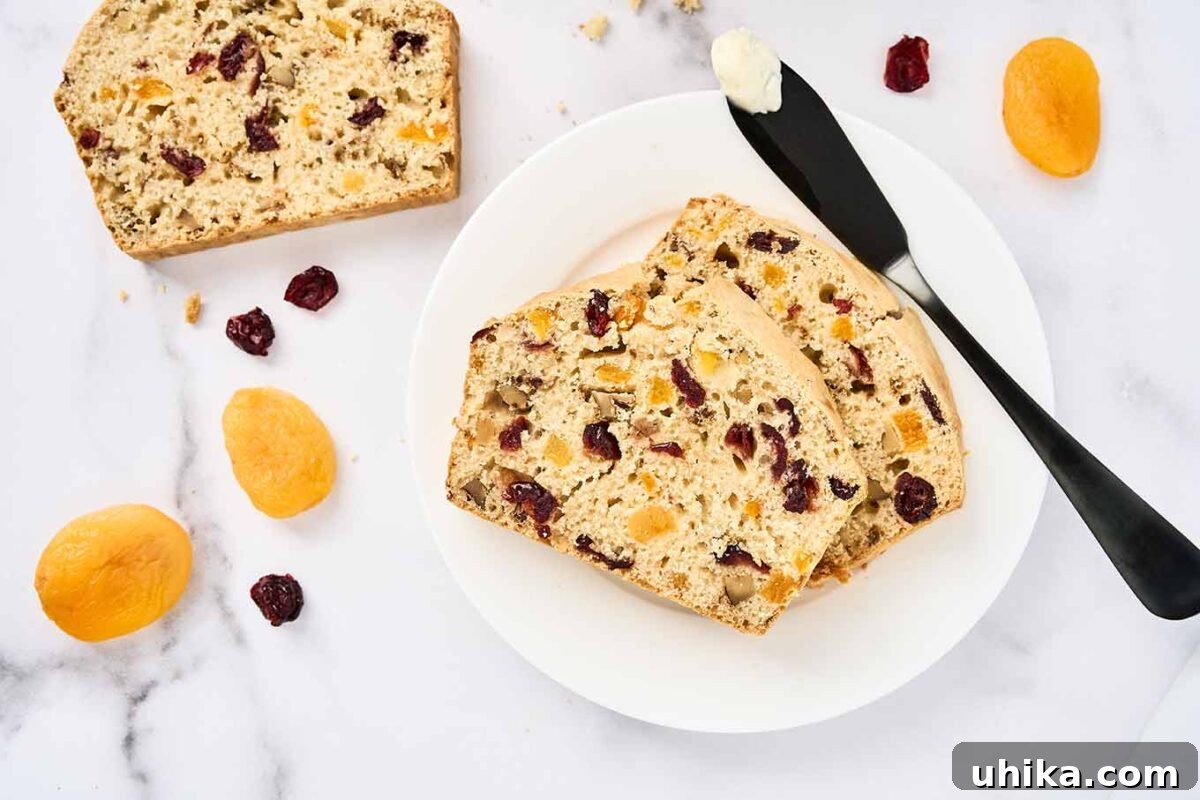 Sun-Kissed Apricot Cranberry Loaf 6 Two slices of apricot cranberry quick bread on a white plate with a buttered knife, surrounded by scattered dried fruits on a marble surface.