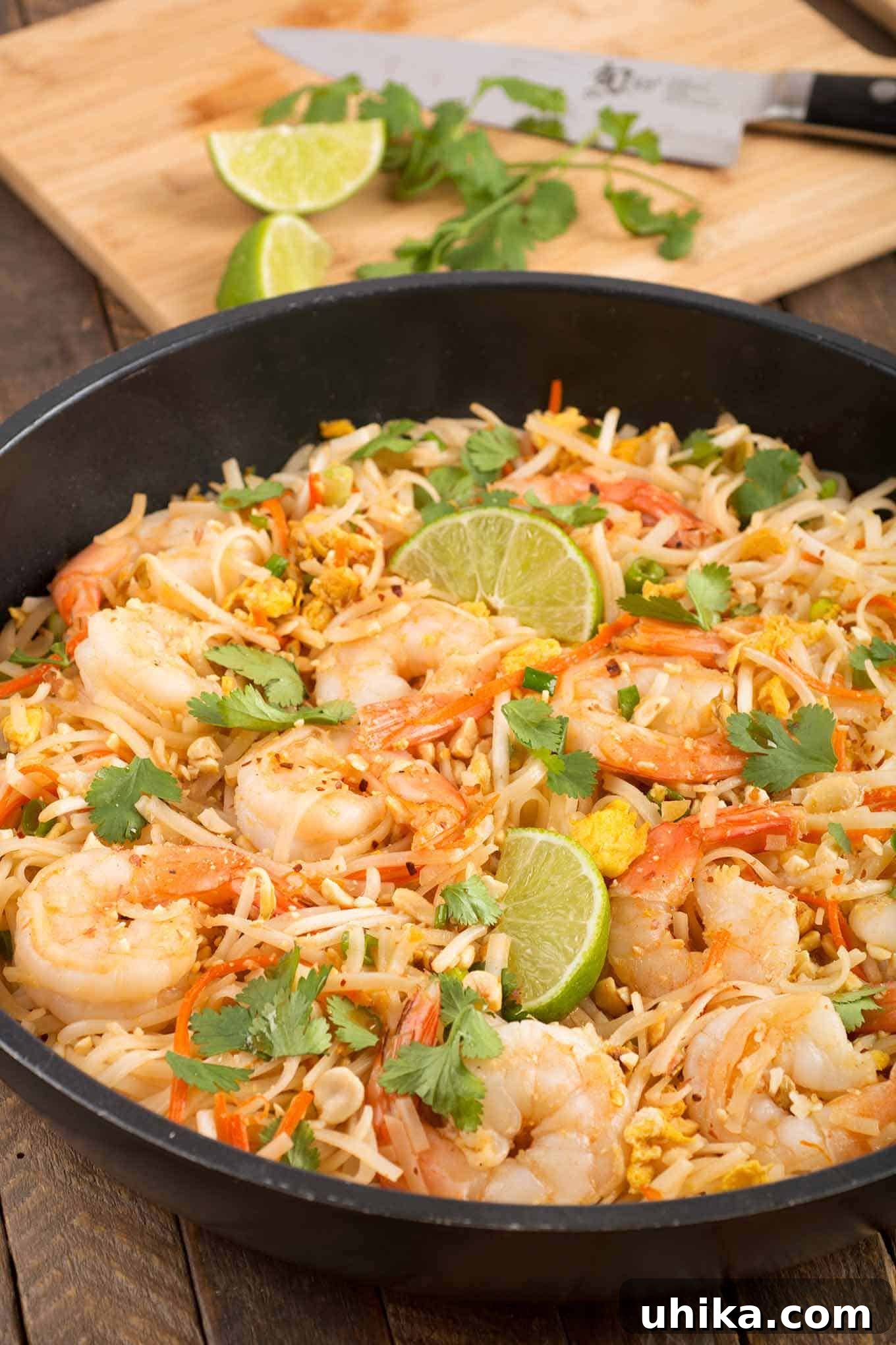 Shrimp Pad Thai noodles in a sauté pan garnished with lime wedges and cilantro, a cutting board with extra lime and cilantro in the background.
