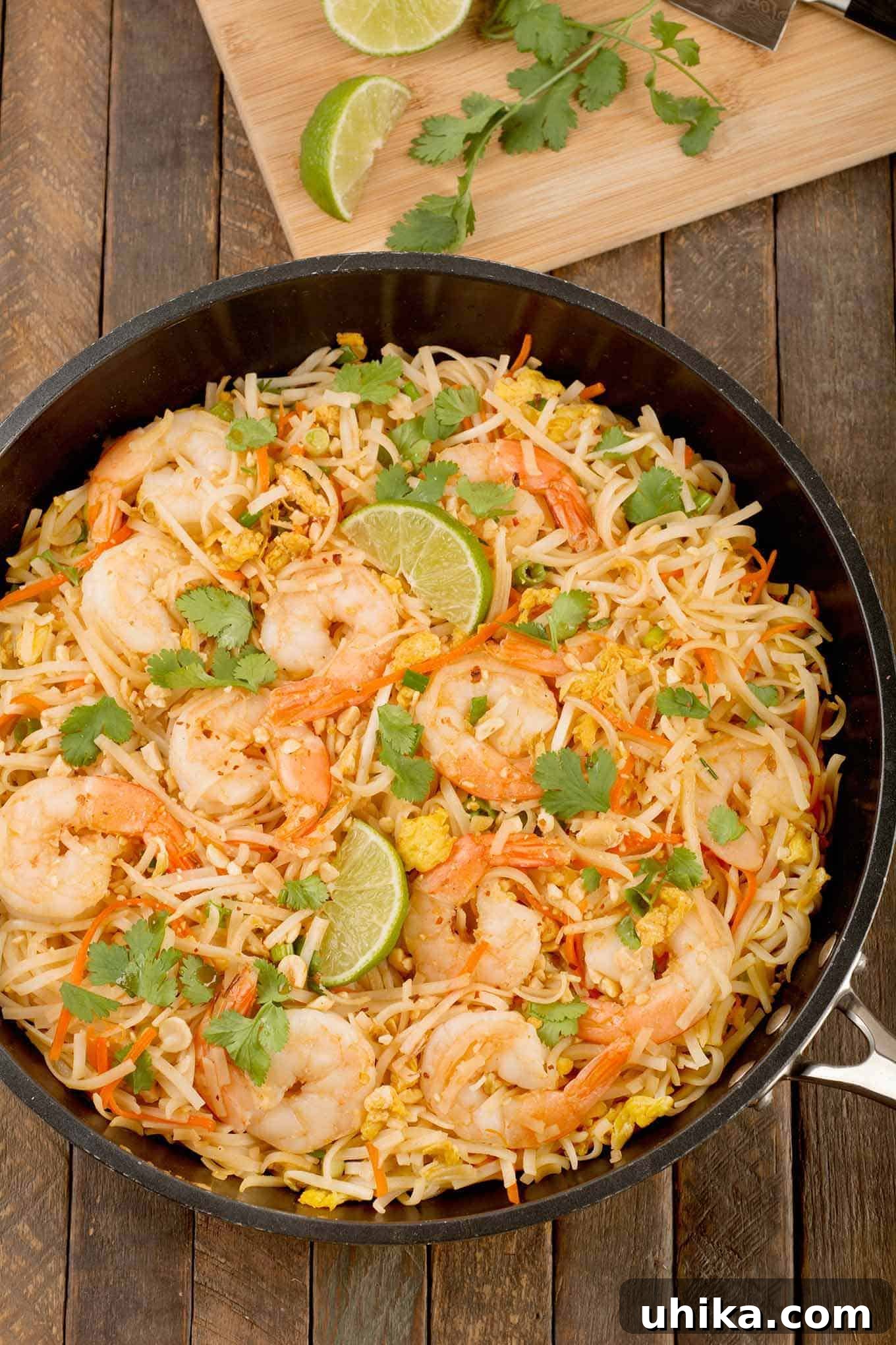 An ovehead view of a sauté pan filled with pad Thai noodles, egg, carrot, bean sprouts, sautéed shrimp, cilantro leaves, and fresh lime.