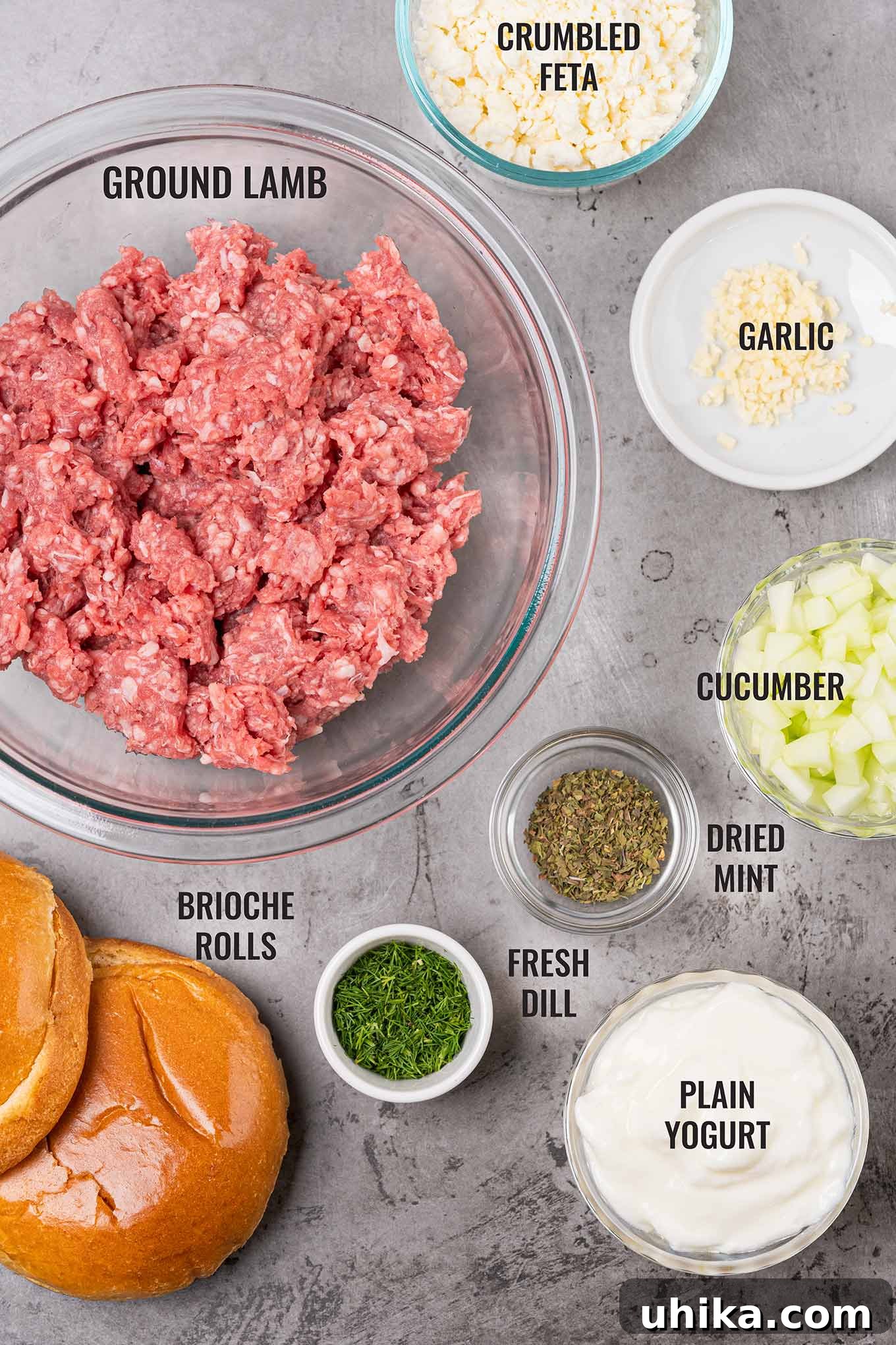 An overhead shot showcasing all the fresh ingredients needed for grilled lamb burgers and homemade tzatziki sauce, including ground lamb, brioche buns, dried mint, cucumber, Greek yogurt, fresh dill, garlic, and tomatoes, neatly arranged on a rustic surface.