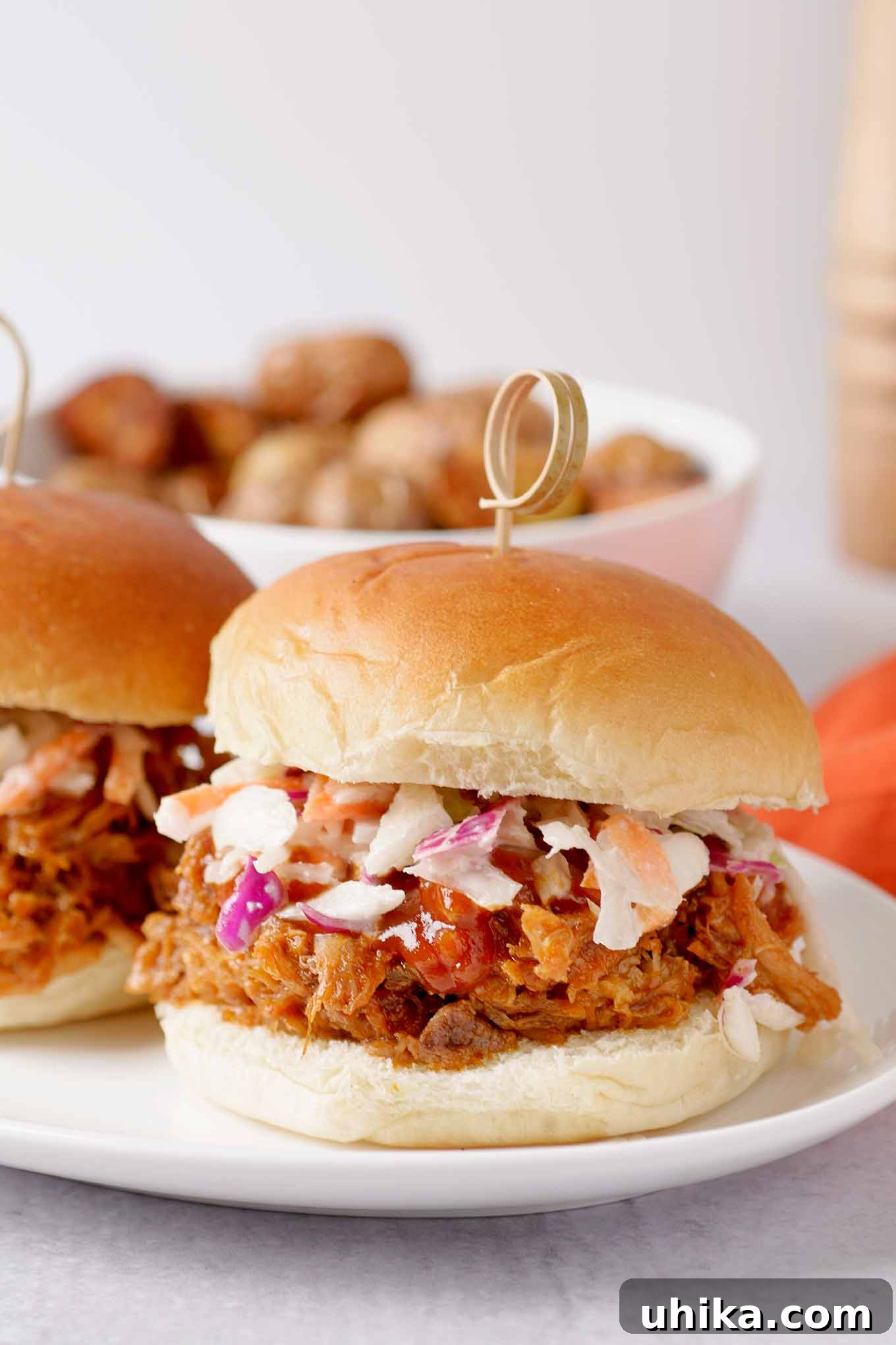 Fall-Apart Tender Oven Pulled Pork 2 A pulled pork sandwich on a soft roll, topped with coleslaw and barbecue sauce, a bowl of roasted potatoes in the background.