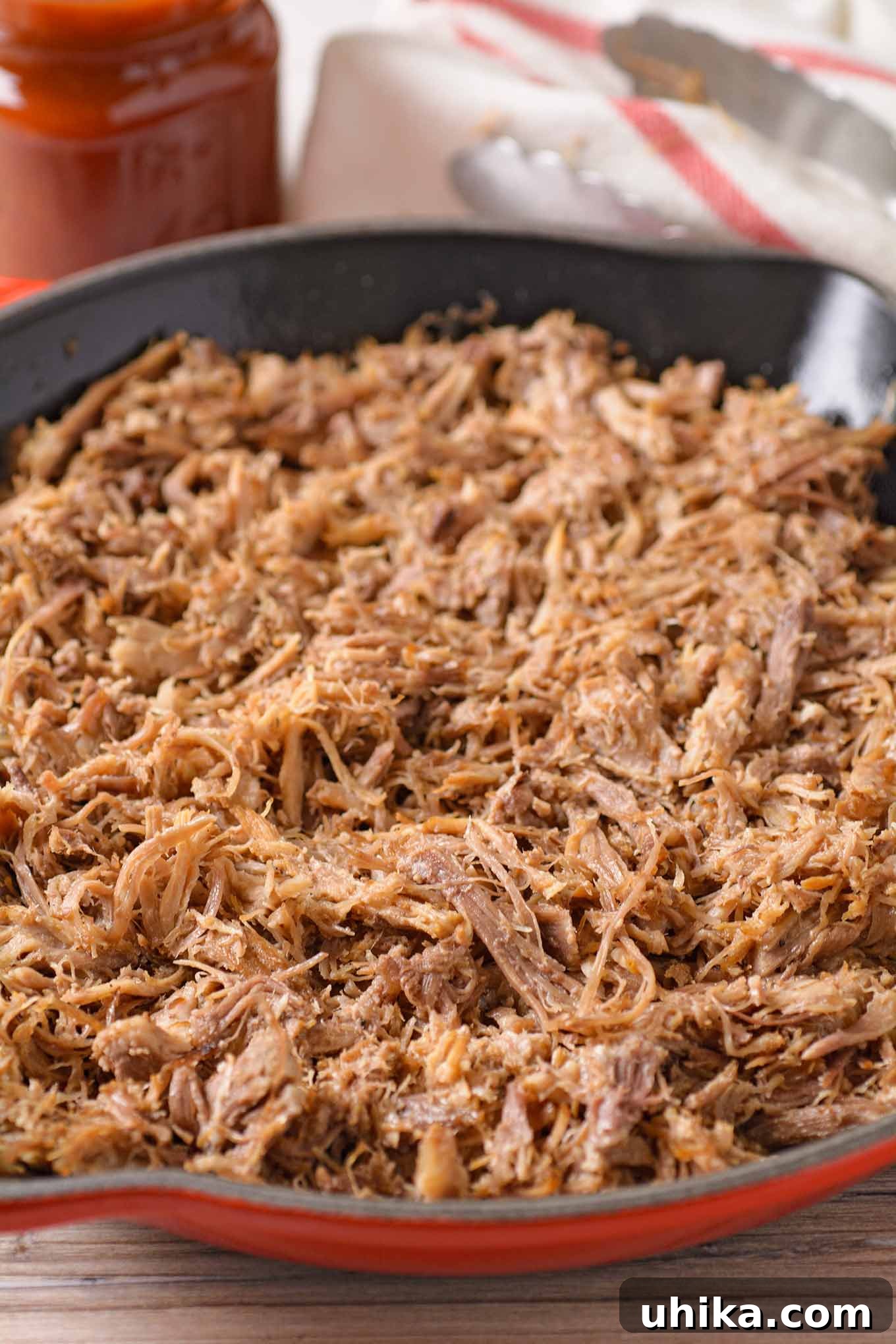 Fall-Apart Tender Oven Pulled Pork 5 A close up of pulled pork in a cast iron skillet before adding sauce, a jar of sauce, kitchen towel, and tongs in the backgroud.