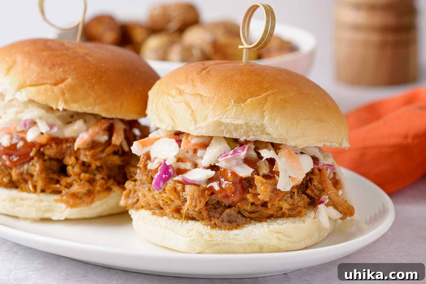 Fall-Apart Tender Oven Pulled Pork 6 A close up of two pulled pork sandwiches on a plate, topped with extra barbecue sauce and homemade coleslaw, secured with a bamboo pick.