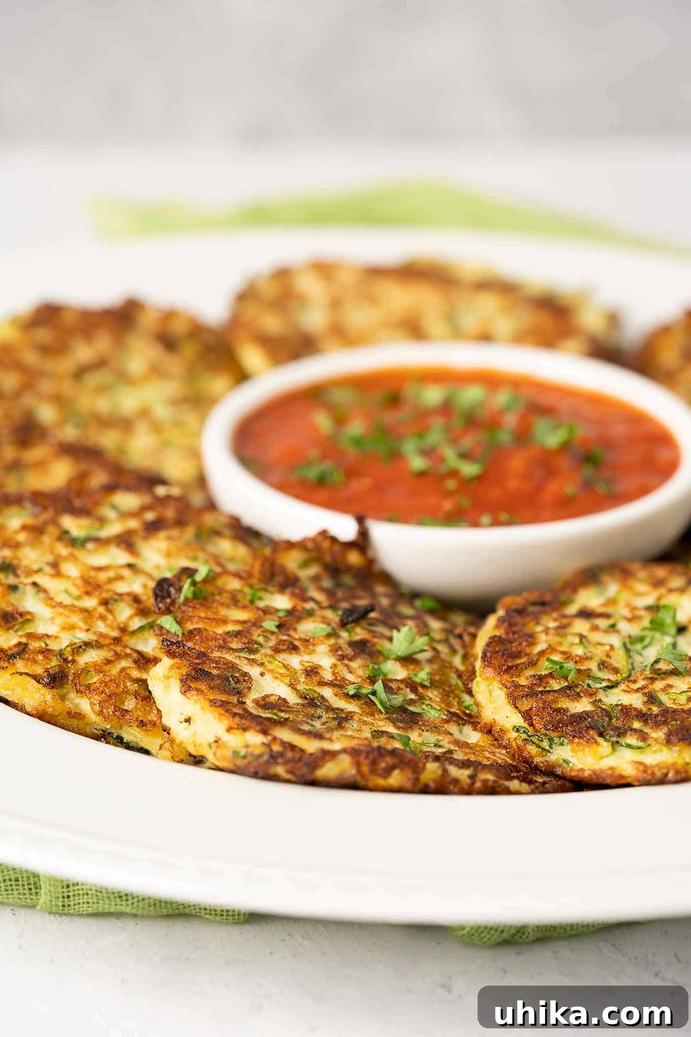 Crispy Parmesan Zucchini Fritters 2 A serving platter of freshly made Parmesan zucchini fritters, golden brown and garnished with fresh parsley, arranged around a small round bowl of marinara dipping sauce.