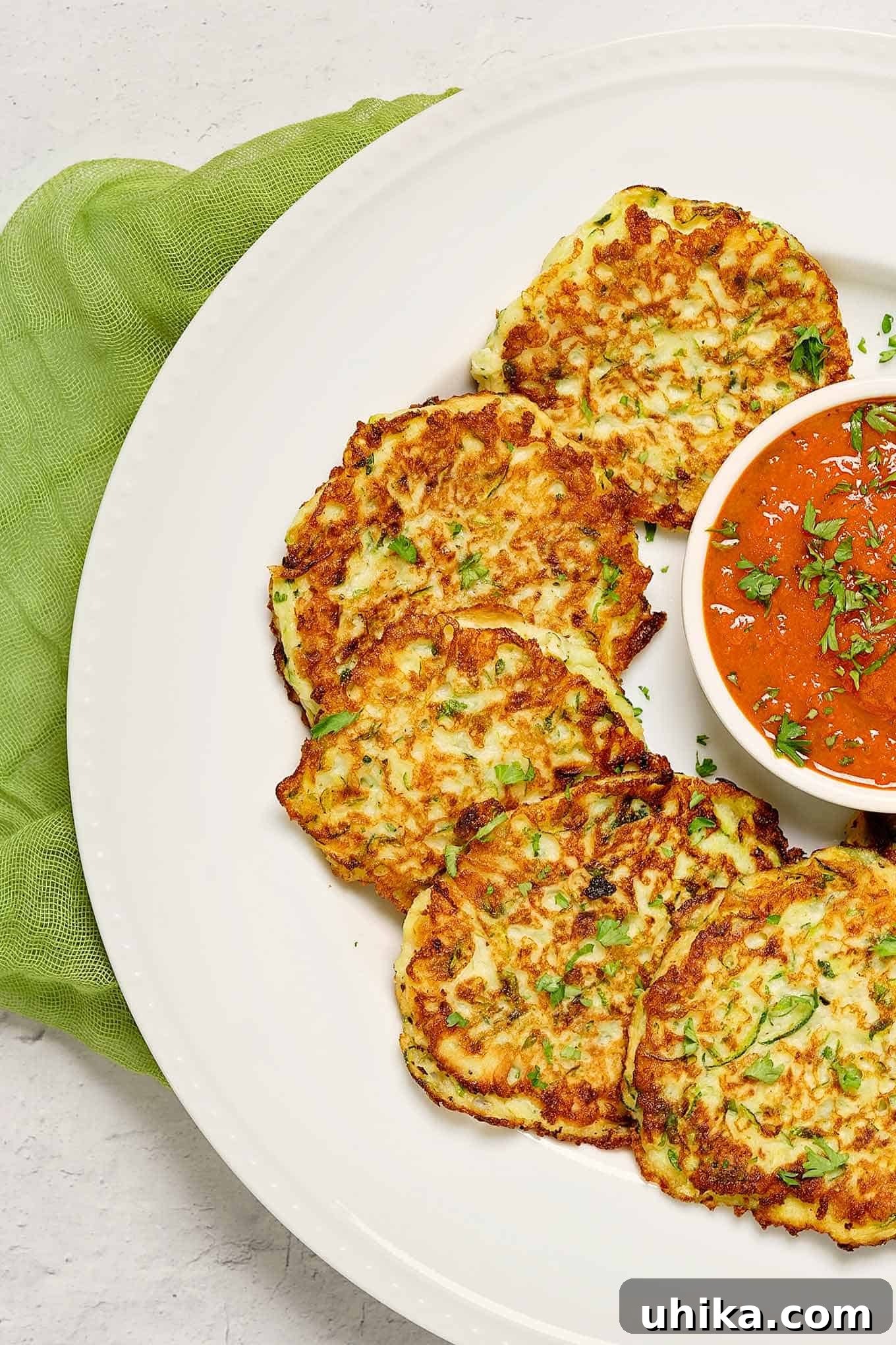 Crispy Parmesan Zucchini Fritters 3 An overhead view of golden brown Parmesan zucchini fritters artfully arranged on a round serving plate, accompanied by a small bowl of marinara sauce and a green linen napkin.