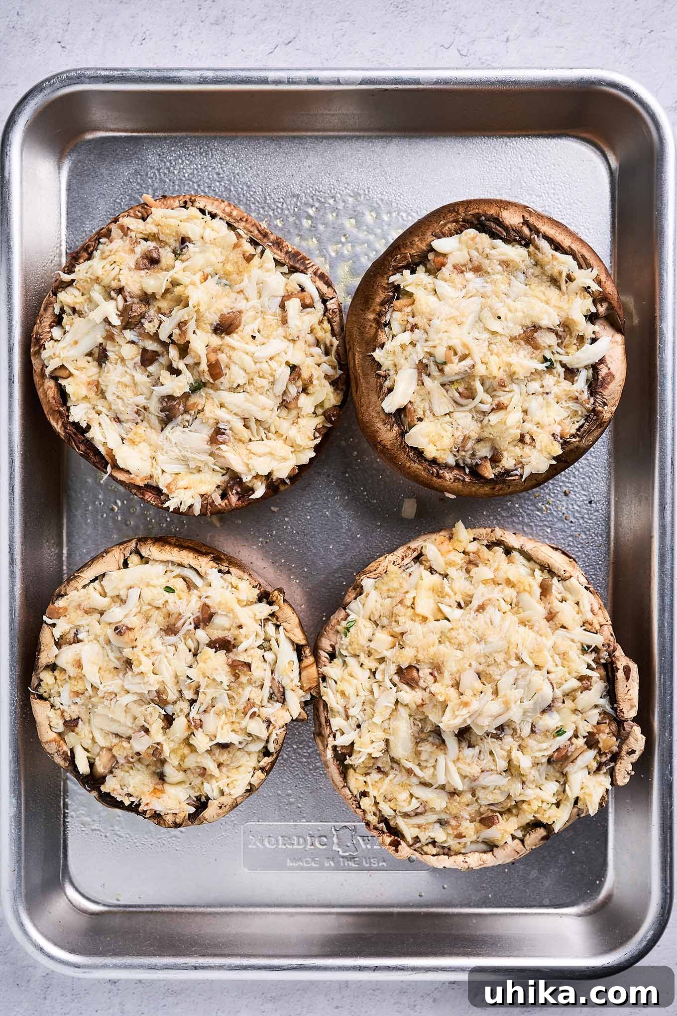 Elegant Crab Stuffed Portobello Caps 5 A baking sheet neatly arranged with four portobello mushroom caps, each generously filled with a savory mixture of lump crabmeat, panko breadcrumbs, and grated Muenster cheese, prepped and ready for the oven.