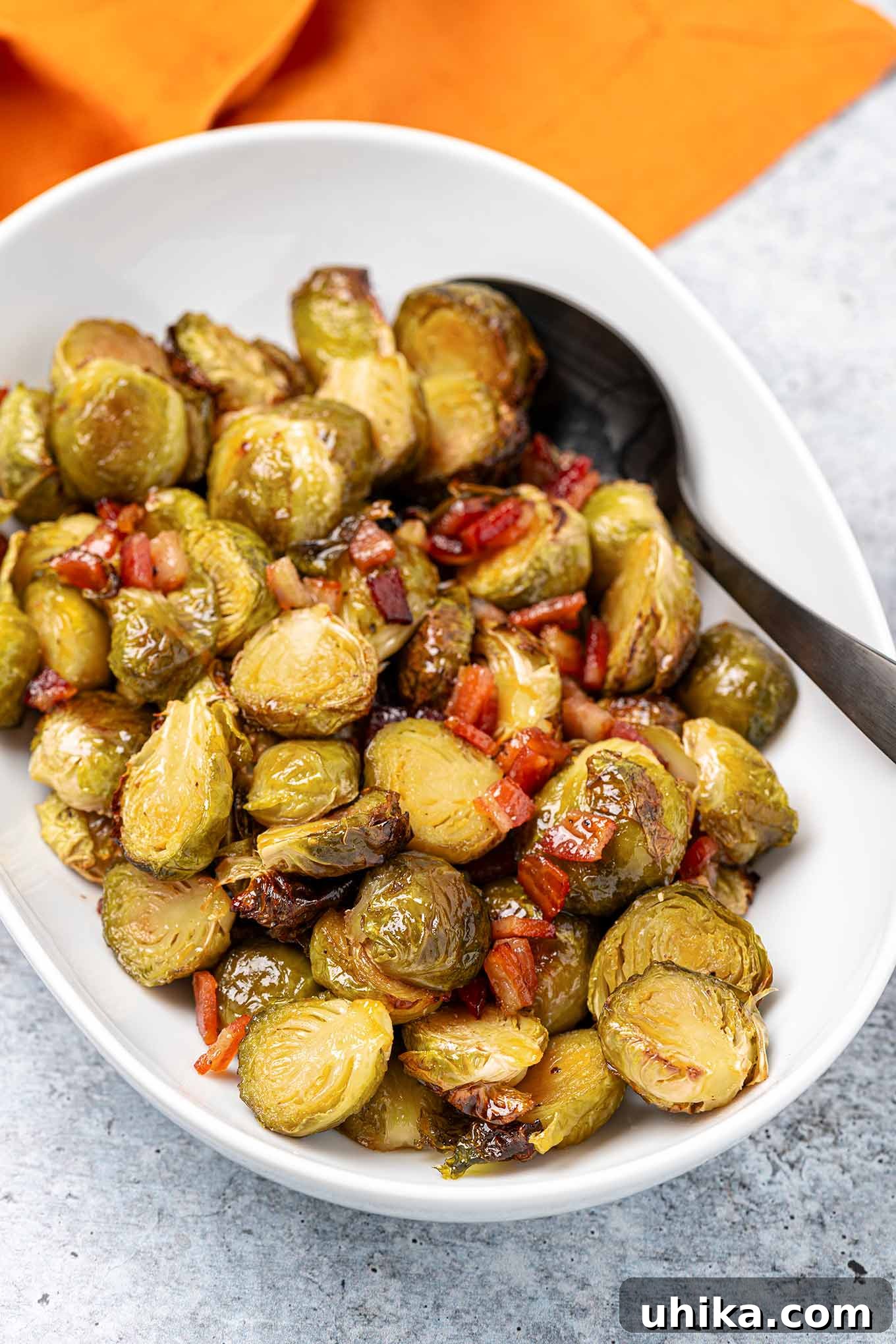 Sticky Maple Bacon Brussels Sprouts 2 An overhead view of Brussels sprouts glazed in maple syrup and butter with bits of thick-cut bacon in a bowl along with a black serving spoon.