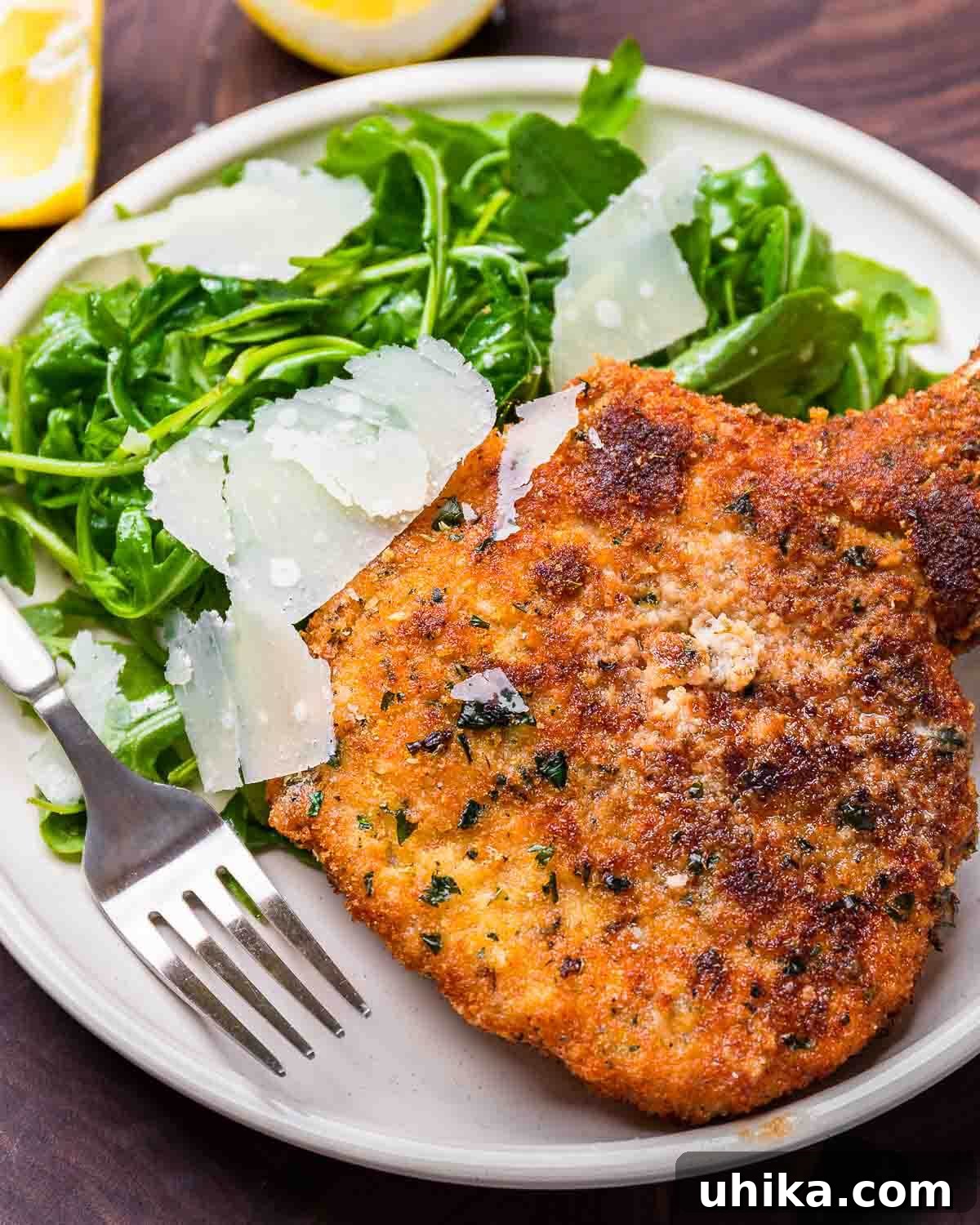 Crispy Milanese Pork Chops 2 Crispy Pork chop Milanese served alongside a fresh arugula salad with shaved parmesan and a lemon wedge on a white plate, ready to be enjoyed.