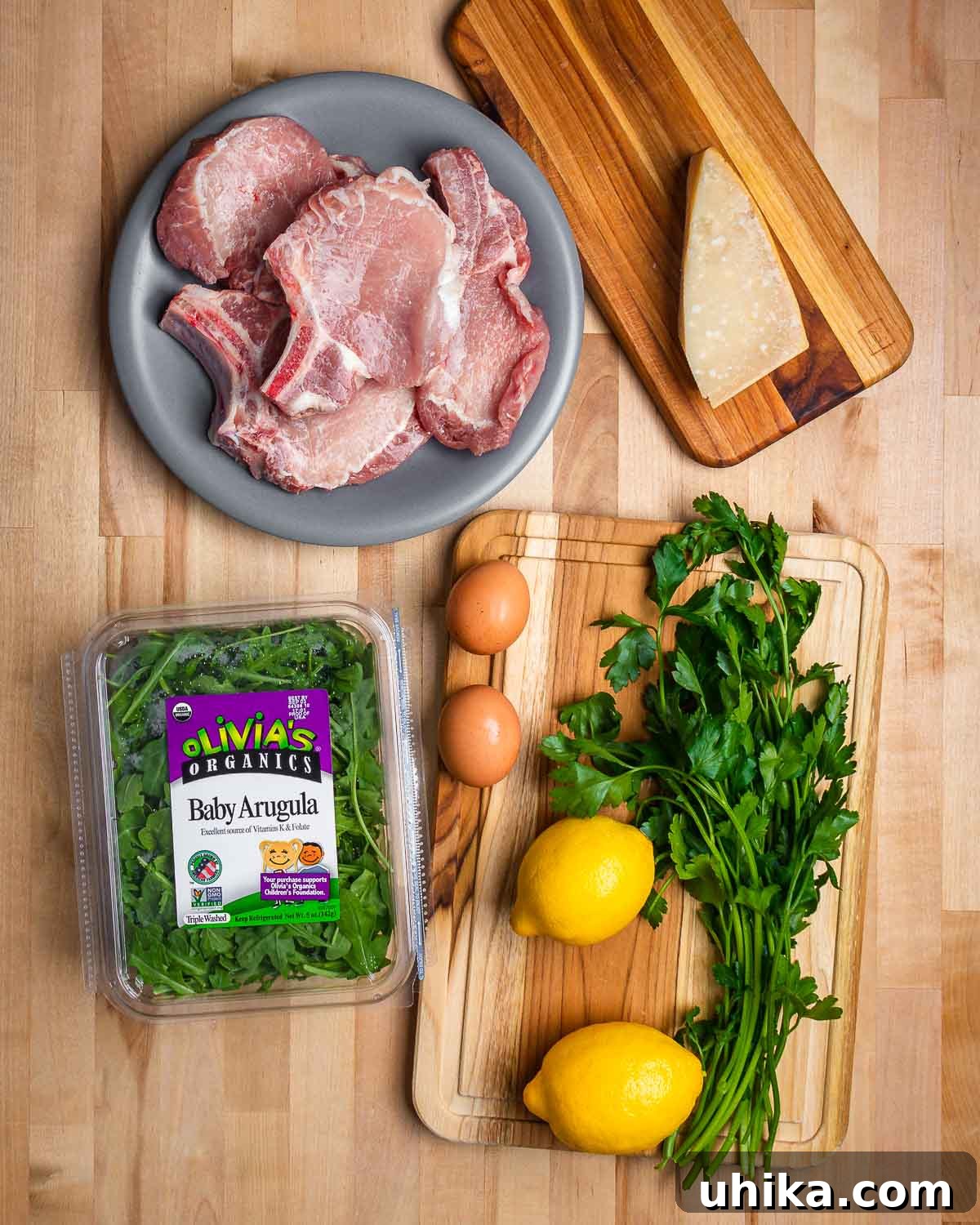 Crispy Milanese Pork Chops 3 Fresh ingredients laid out for Pork Milanese recipe: raw pork chops, a block of Parmigiano Reggiano, a bowl of baby arugula, eggs, lemons, and fresh parsley sprigs.