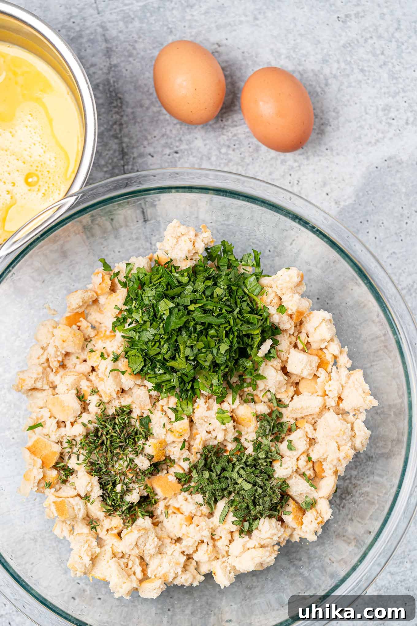 Heirloom Bread Stuffing 4 Preparing bread stuffing, one mixing bowl with moistened bread cubes, fresh thyme, parsley, and sage, beaten eggs in a separate bowl to the side.
