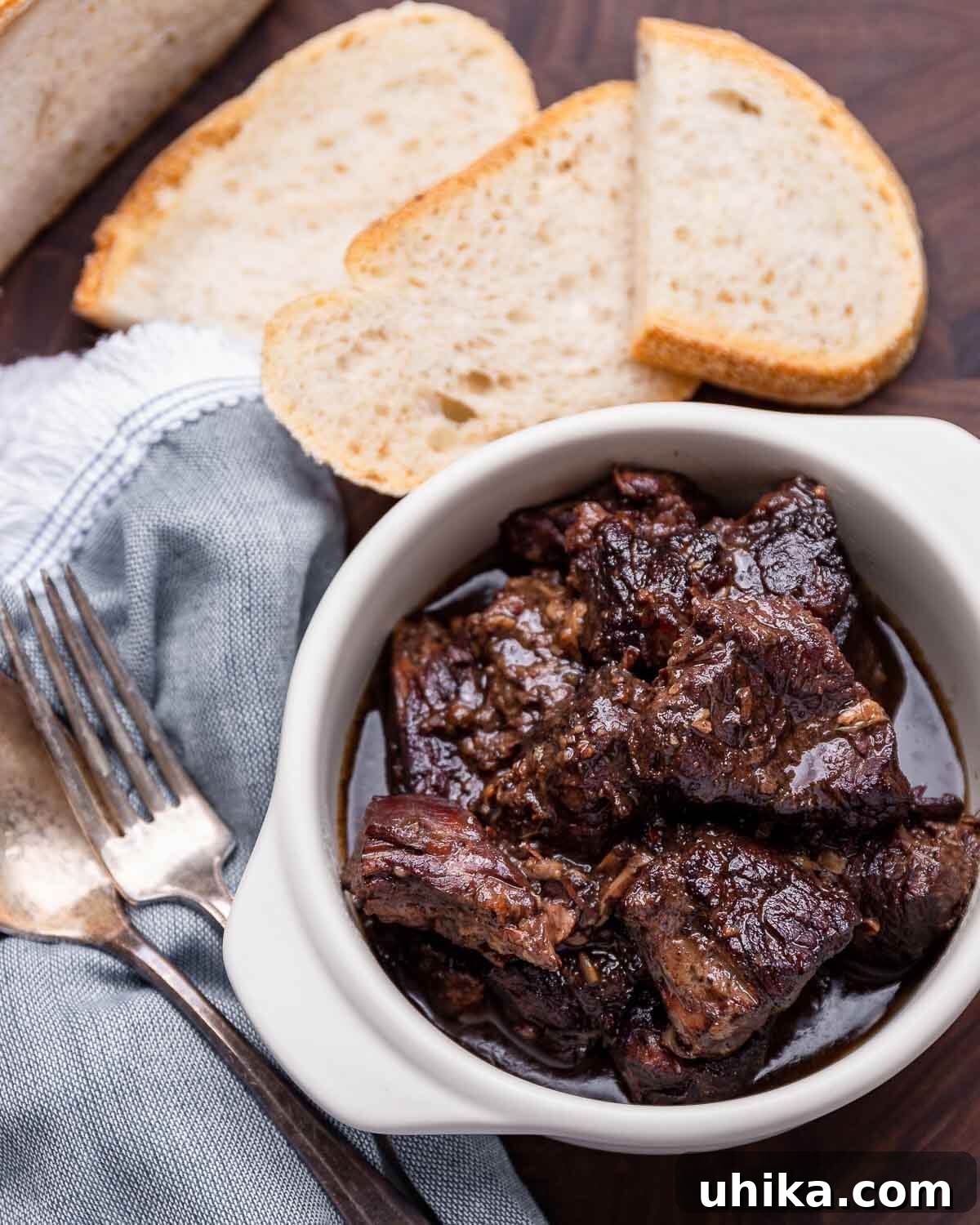 Tuscan Peppery Beef 2 A rustic bowl of Peposo, a Tuscan beef stew, garnished with fresh parsley and served alongside crusty bread. A blue napkin, fork, and spoon are neatly arranged on the side.