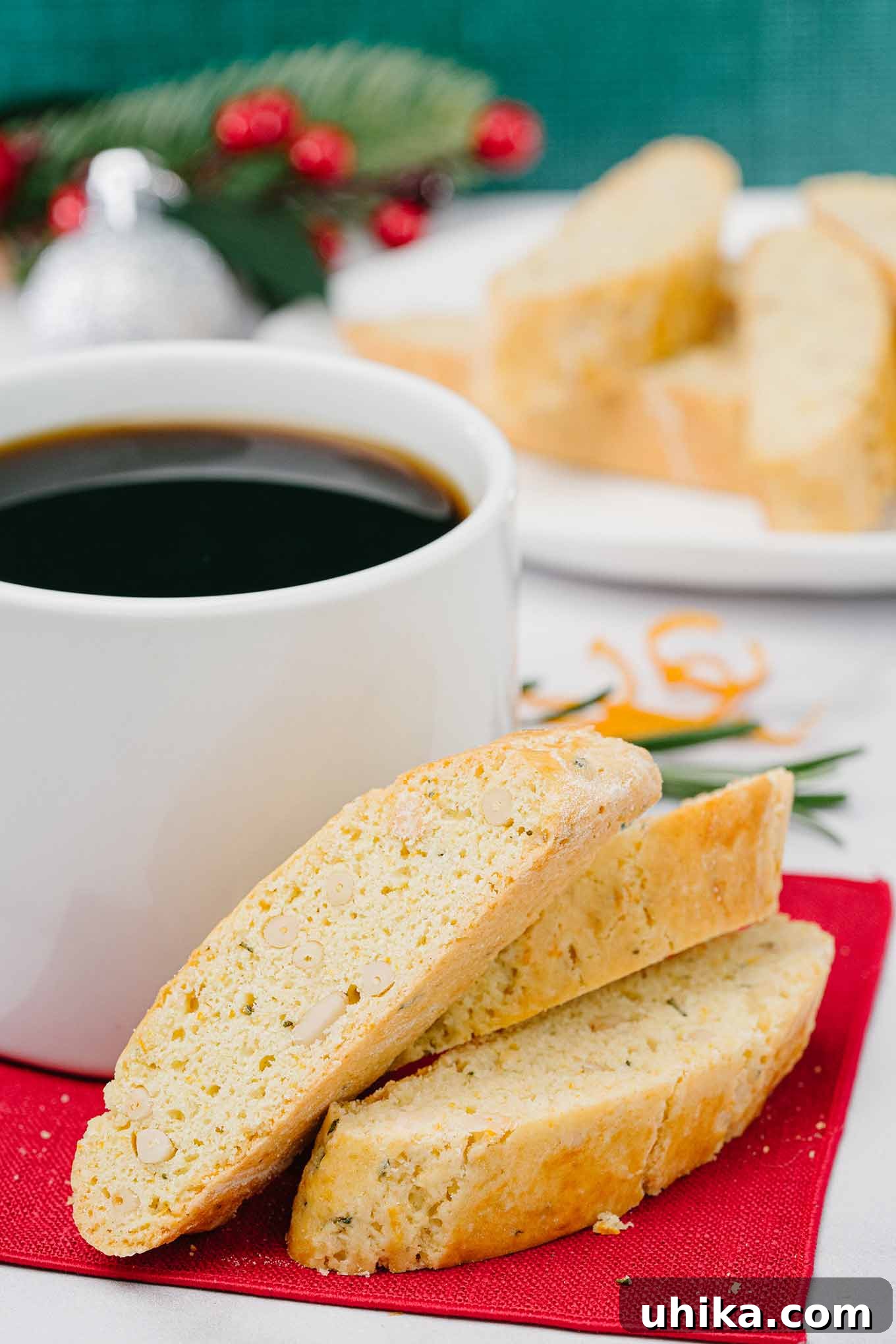Zesty Butter Biscotti: A Double Treat 2 Buttery orange biscotti with pine nuts and rosemary on a red napkin, beside a coffee mug, with more biscotti and festive decor in the background.