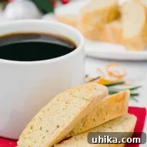Buttery orange biscotti with pine nuts and rosemary on a red napkin, beside a coffee mug, with more biscotti and festive decor in the background.