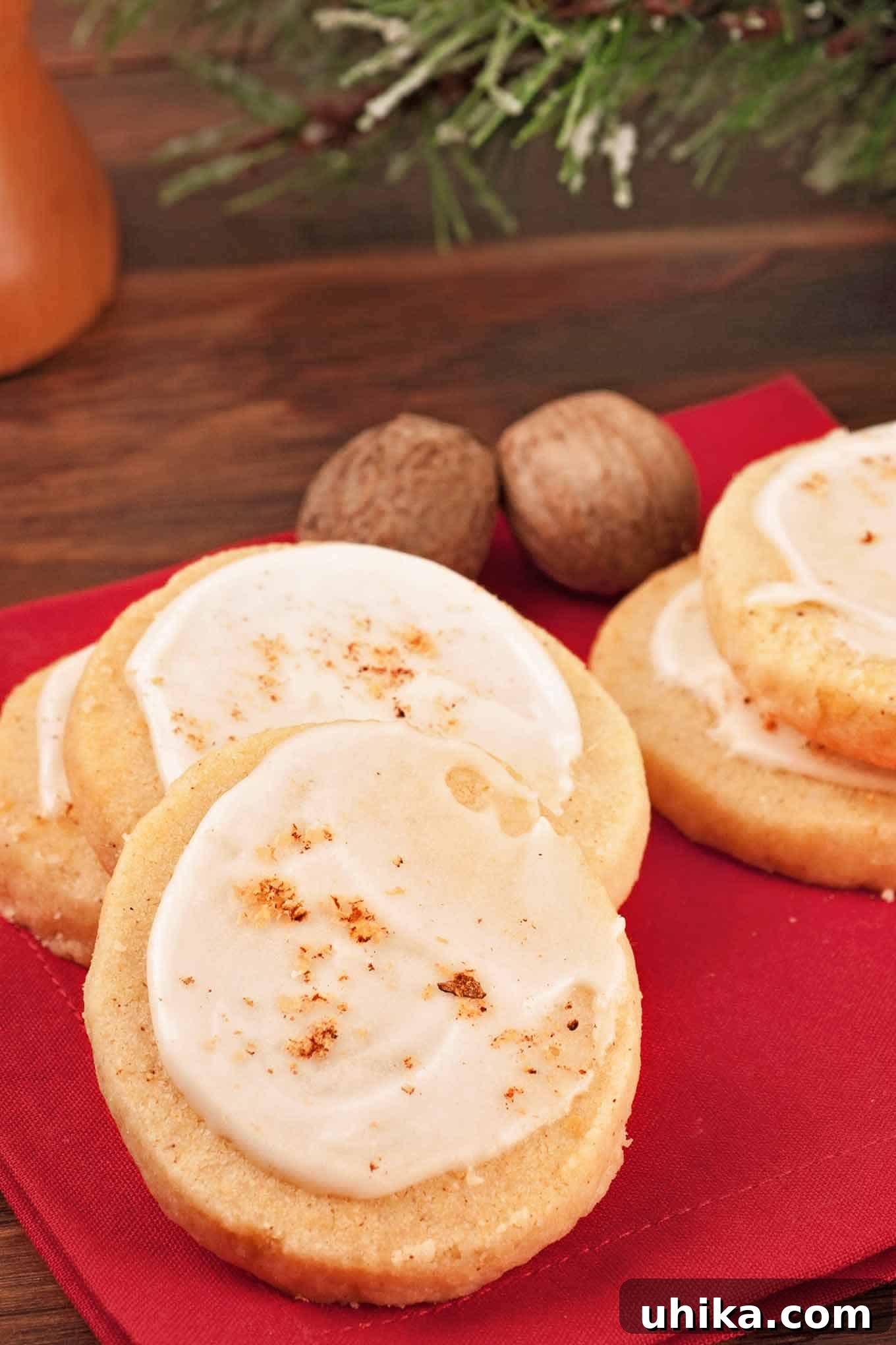 Eggnog Refrigerator Cookies 2 Eggnog cookies topped with rum icing and a sprinkle of nutmeg arranged on a red napkin, with whole nutmeg pods in the background.