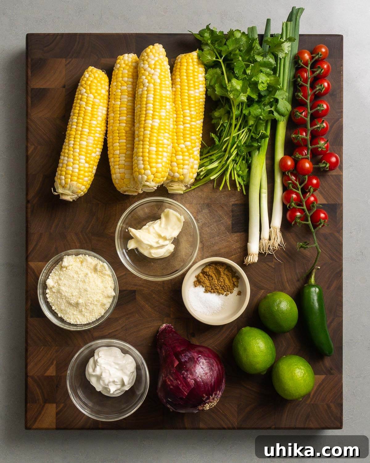 Esquites Corn Salad 3 A colorful array of ingredients laid out on a table, including corn cobs, fresh cilantro, green onions, halved cherry tomatoes, crumbled cotija cheese, creamy mayonnaise, tangy sour cream, bright red onion, juicy limes, and a spicy jalapeño pepper.
