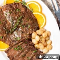 A serving platter of partially carved beef brisket garnished with oranges, pearl onions, fresh rosemary and thyme.