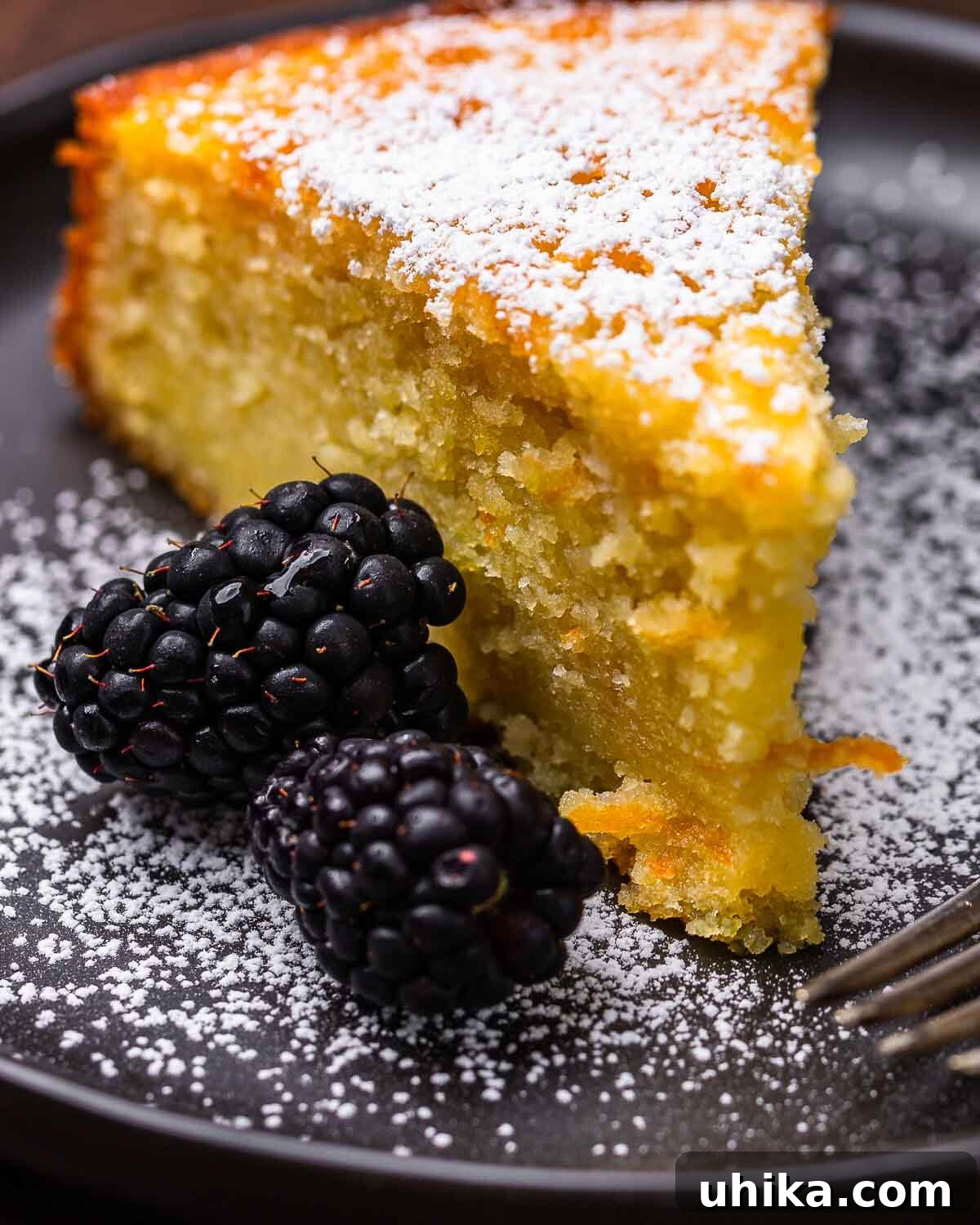 Piece of orange olive cake in plate with powdered sugar and blackberries.