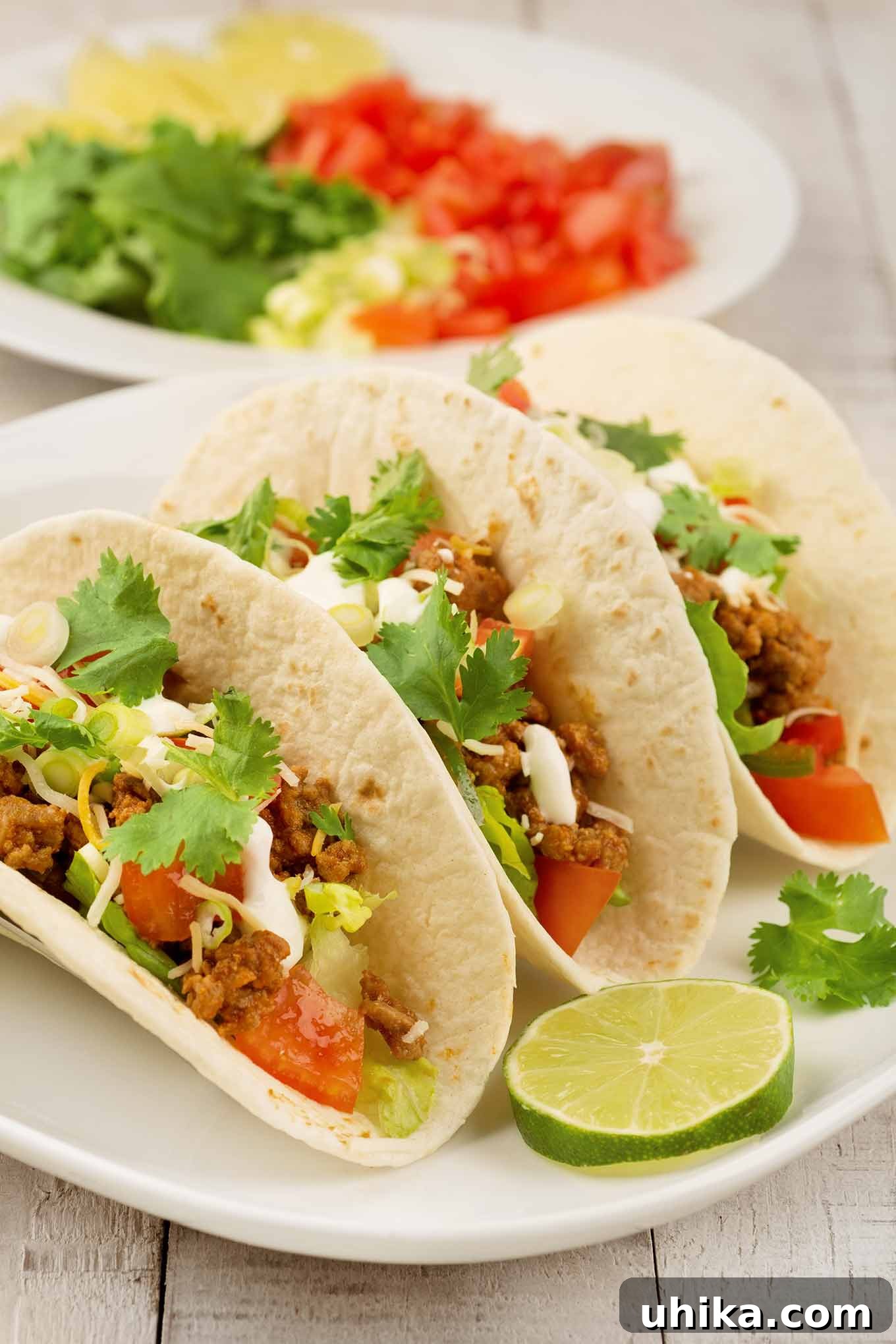 Zesty Turkey Tacos 2 Three soft ground turkey tacos on a plate garnished with lime and cilantro, with a plate of taco toppings in the background, showcasing a delicious, healthy meal.