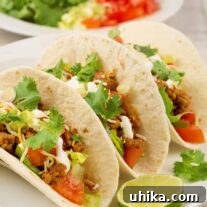 Three soft ground turkey tacos on a plate garnished with lime and cilantro, a plate filled with taco toppings in the background.