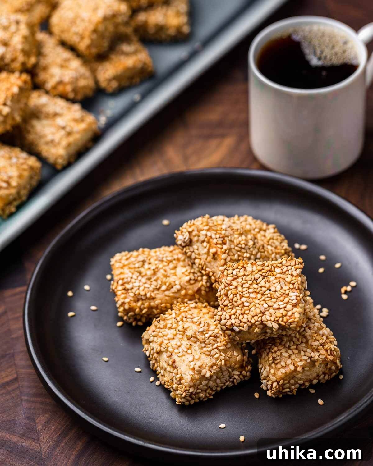 Crispy Sesame Delights 2 Reginelle cookies arranged on a black plate with a cup of coffee in the background, showcasing their golden-brown hue and sesame seed coating.