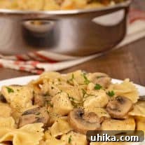 A serving plate of farfalle combined with mushrooms and bite-sized pieces of chicken breasts in a creamy marsala sauce, a sauté pan with more in the background.