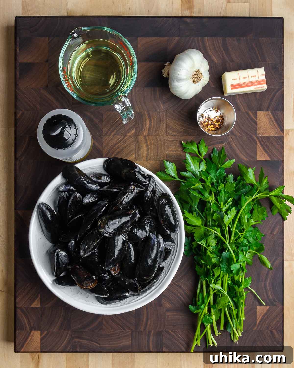 Wine-Infused Mussels 3 An organized arrangement of fresh ingredients including a bottle of white wine, whole garlic cloves, butter sticks, a bottle of olive oil, a small bowl of chili flakes, a bowl of raw mussels, and a bunch of fresh parsley on a kitchen counter, ready for preparation.