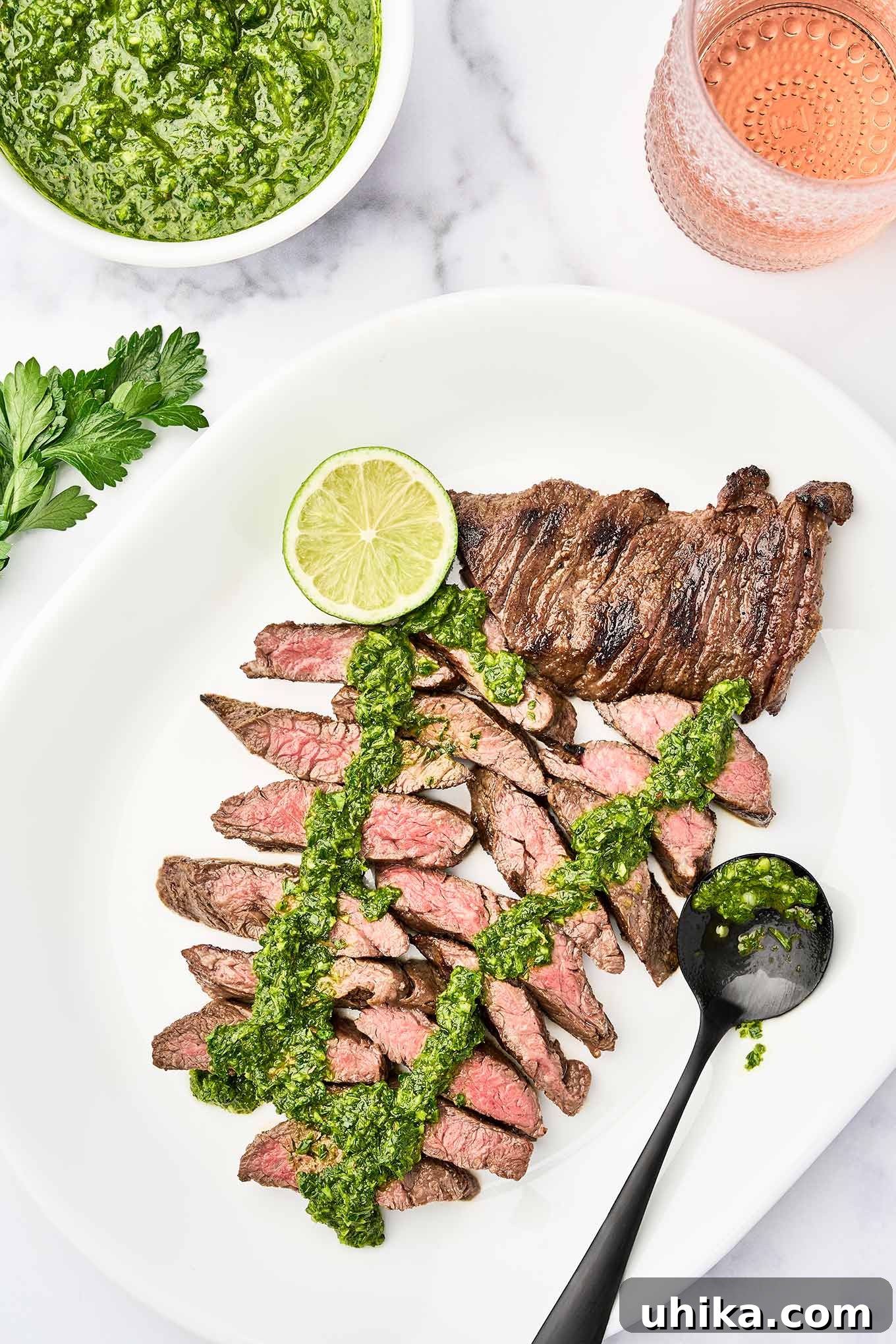 Sizzling Steak, Vibrant Chimichurri 2 A white serving platter with sliced, medium-rare, grilled skirt steak, topped with chimichurri sauce, garnished with a lime slice, a bowl of chimichurri and glass of rosé in the background.