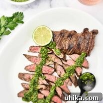 A white serving platter with sliced, medium-rare, grilled skirt steak, topped with chimichurri sauce, garnished with a lime slice, a bowl of chimichurri and glass of rosé in the background.