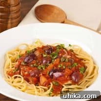 Plate of Pasta alla Puttanesca with spaghetti topped with tomato sauce, kalamata olives, capers, onions, and garnished with chopped parsley.