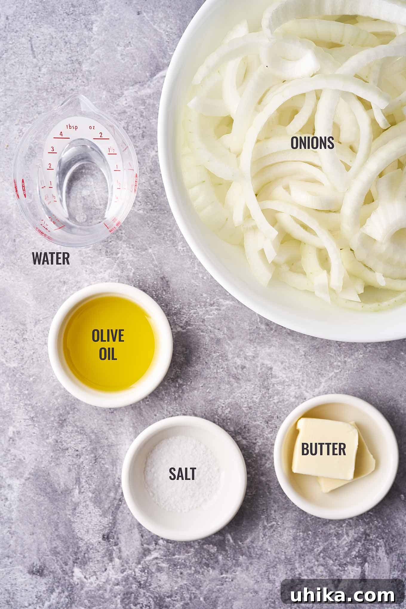 An overhead view of ingredients for caramelized onions, neatly arranged: thinly sliced yellow onions, olive oil, butter, and a small dish of salt, along with a container of water.
