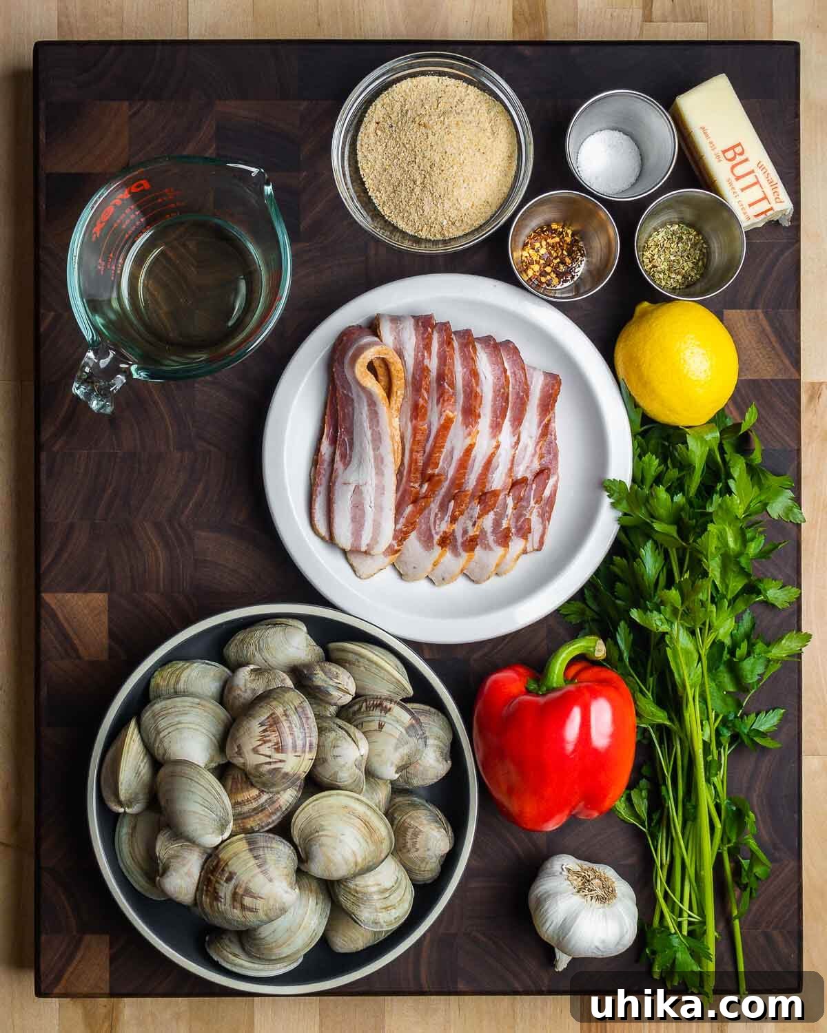 Baked Clams Royale 3 A vibrant display of fresh ingredients for Clams Casino, including pristine Littleneck clams, smoky bacon, bright red bell pepper, aromatic garlic cloves, fresh parsley, lemons, golden breadcrumbs, dried oregano, red pepper flakes, butter, and a bottle of white wine. Note: A generous amount of bacon is shown, but only a few strips are needed for this recipe.